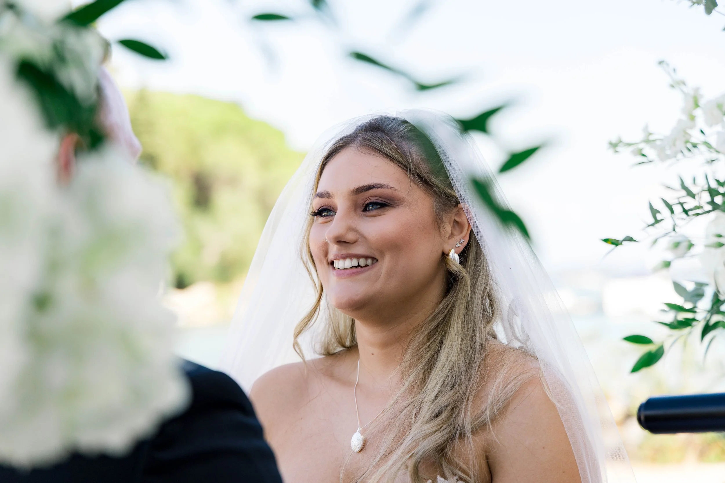 A smiling bride with blonde hair and blue eyes, wearing a white wedding dress, veil, pearl necklace, and earrings, during an outdoor wedding ceremony with greenery in the background.