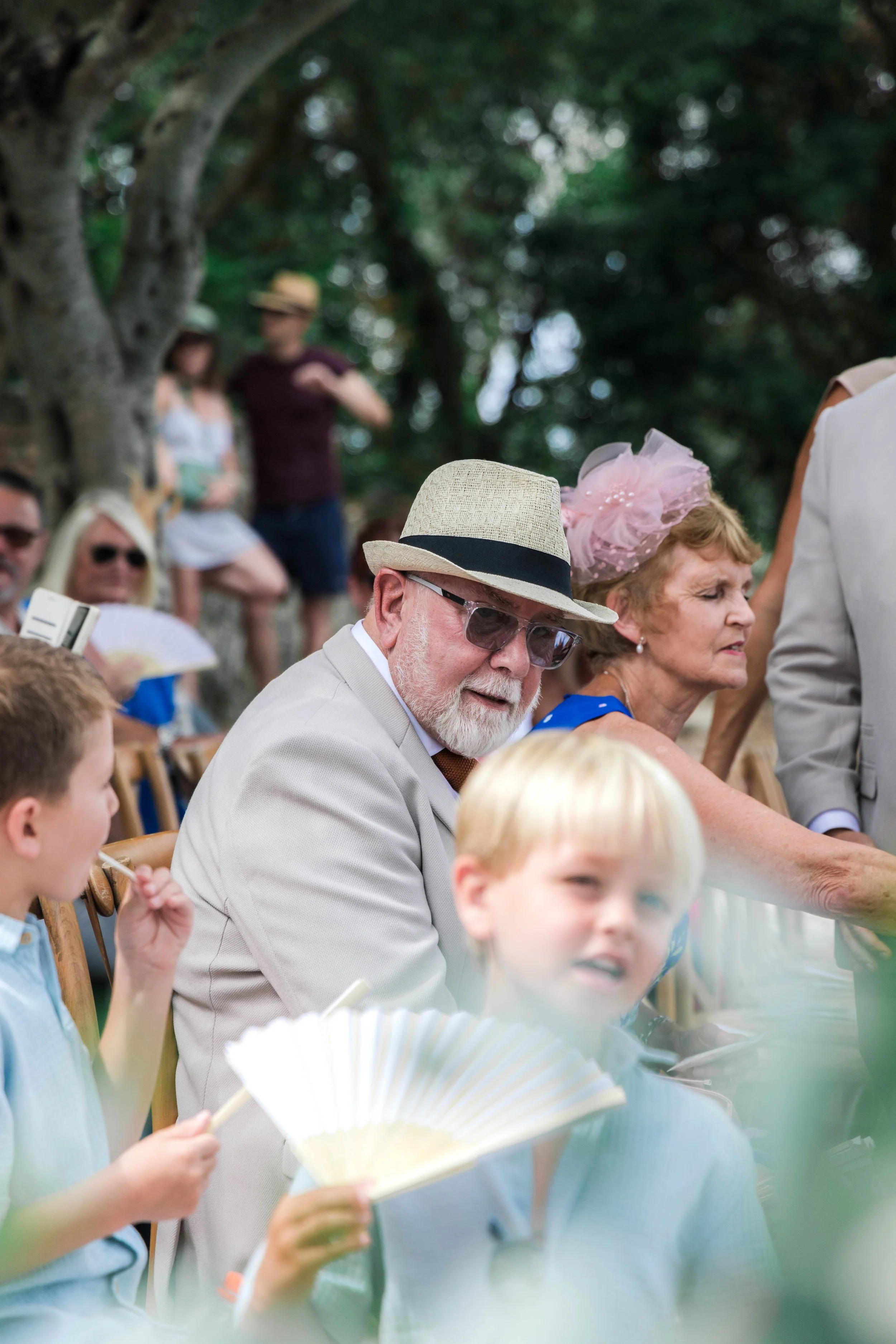 A group of people sitting outdoors enjoying an event, including a man wearing a straw hat and sunglasses, and a woman wearing a pink hat with netting. Some people are holding fans. The scene is shaded by a large tree.