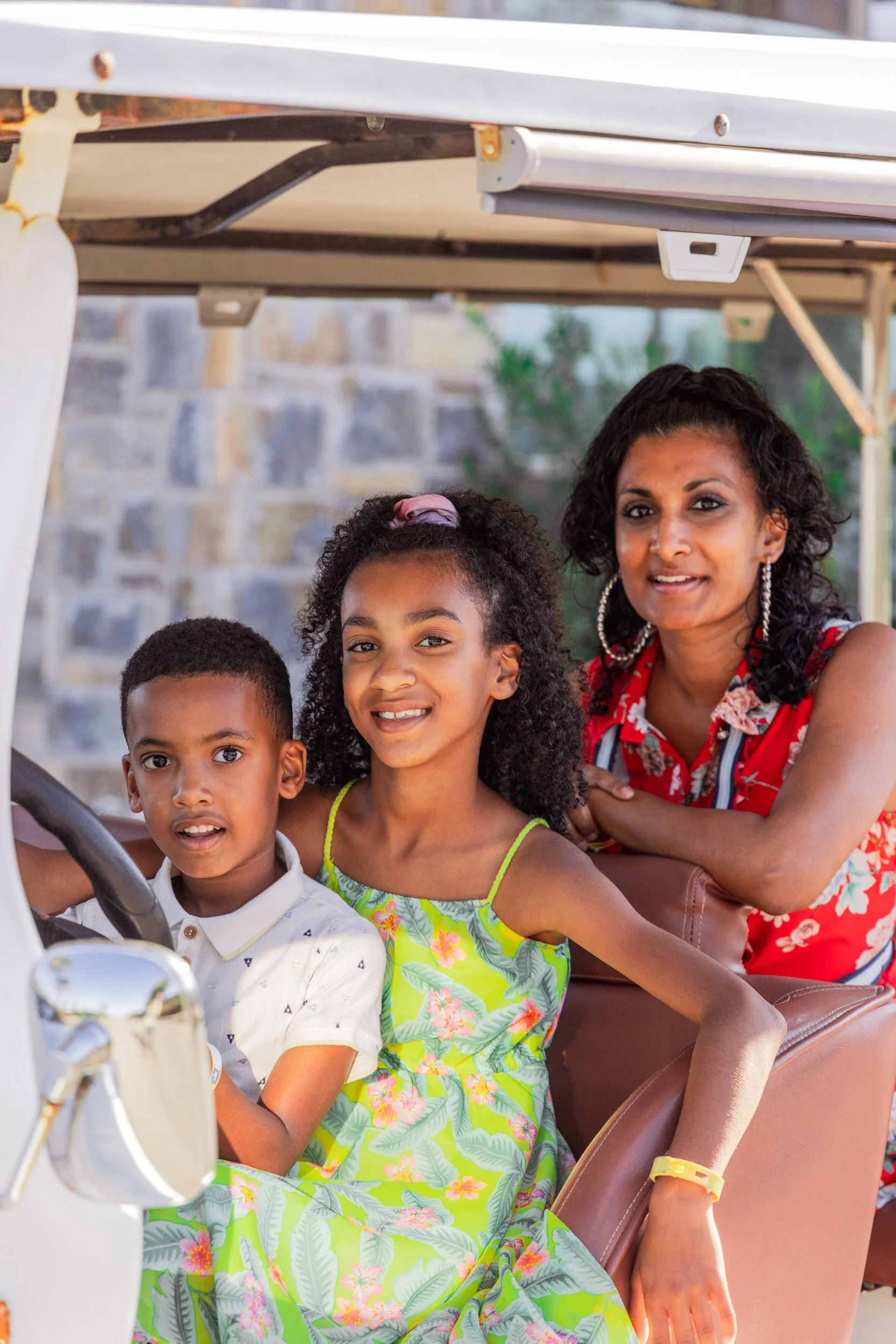 A woman with dark curly hair and earrings, sitting next to two children in a golf cart, with a stone wall in the background.