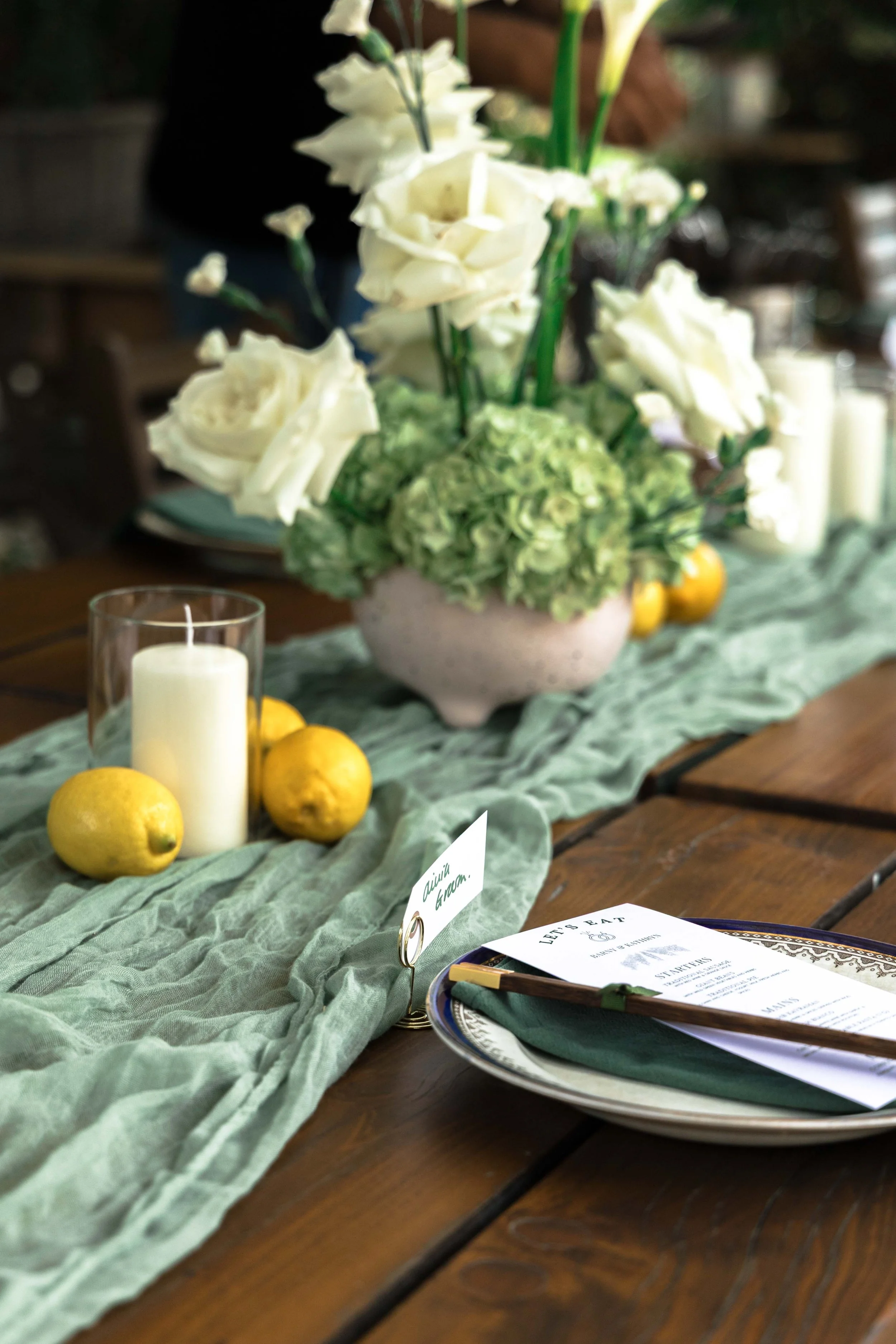 Table centerpiece with white flowers in a pink vase, yellow lemons, a glass candle holder with a white candle, a plate with a green napkin, a menu, and a small card that says "With Grace" on a wooden table with a green table runner.