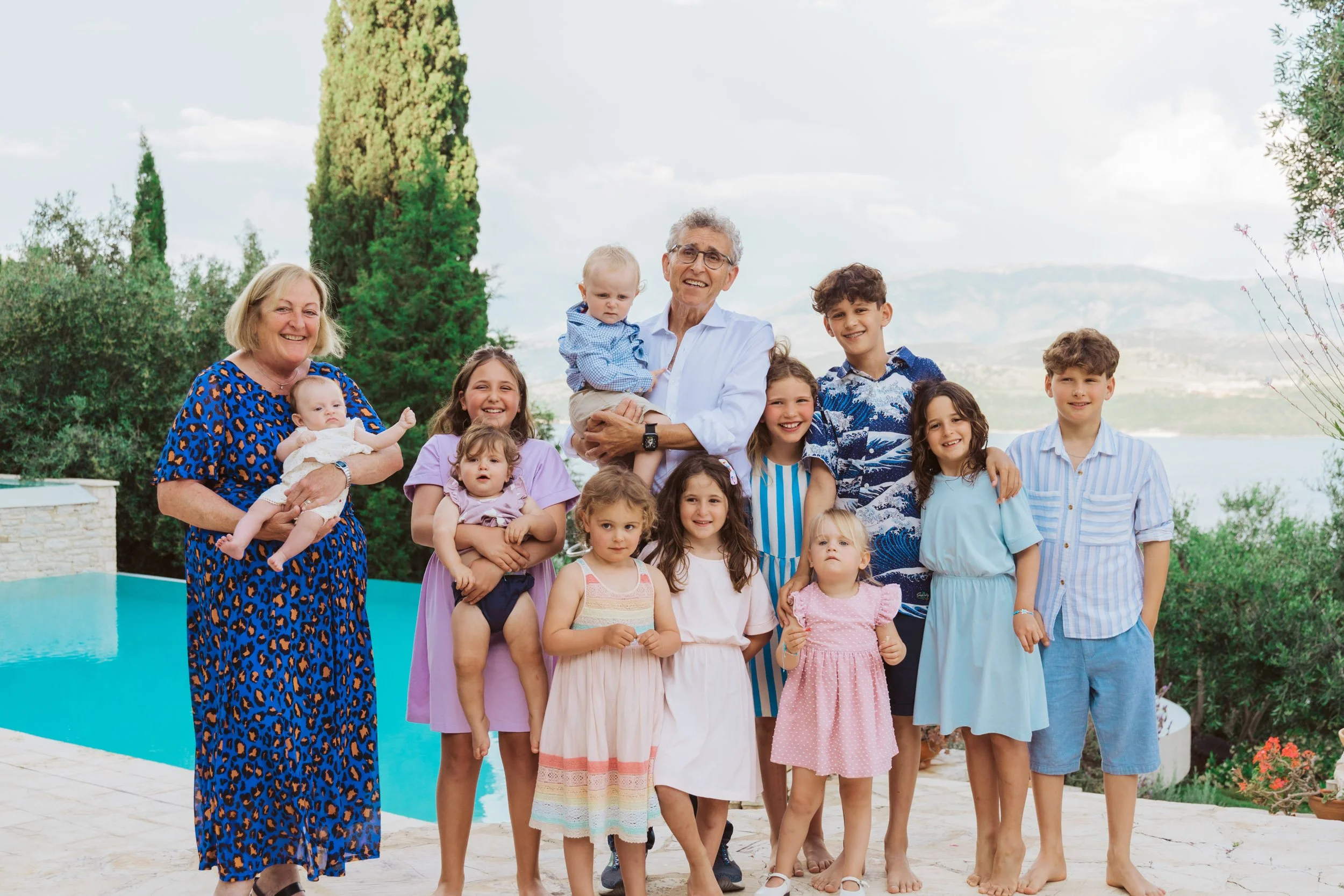 A family of sixteen people, including grandparents, parents, and children, posing outdoors near a swimming pool with trees and mountains in the background.