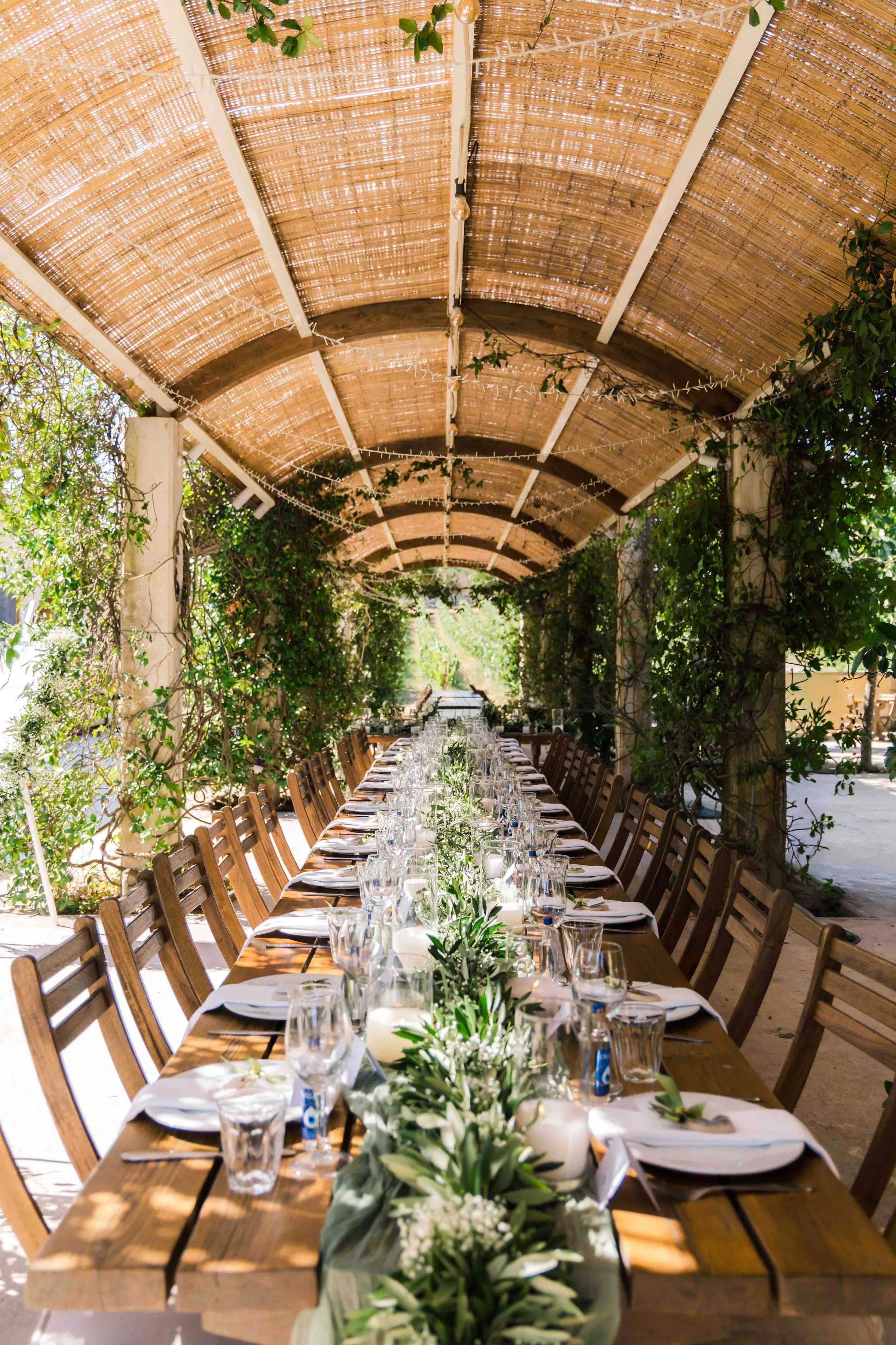 Long outdoor dining table set for a formal event under a wooden, bamboo-covered pergola surrounded by greenery.