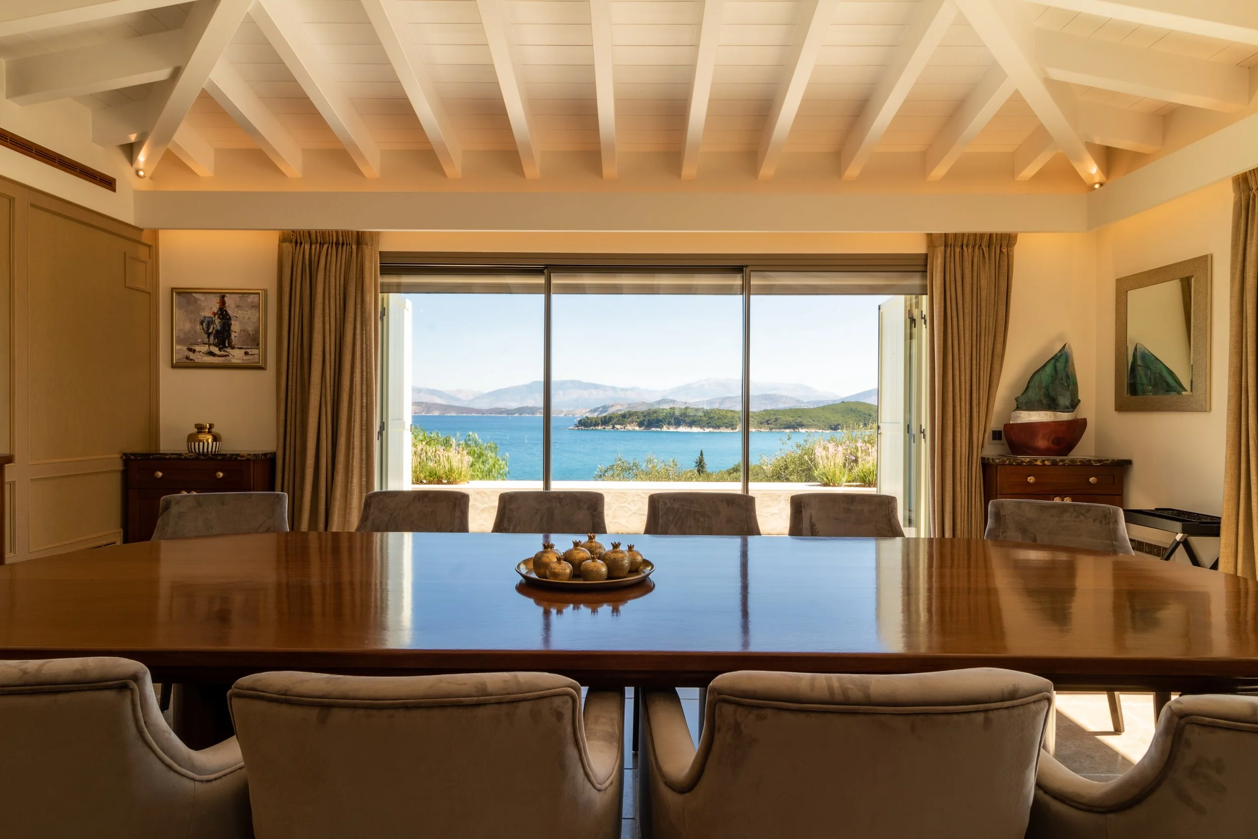 Room with a large wooden dining table and upholstered chairs, overlooking a scenic view of a lake and mountains through large sliding glass doors, with beige curtains and decorative artwork.
