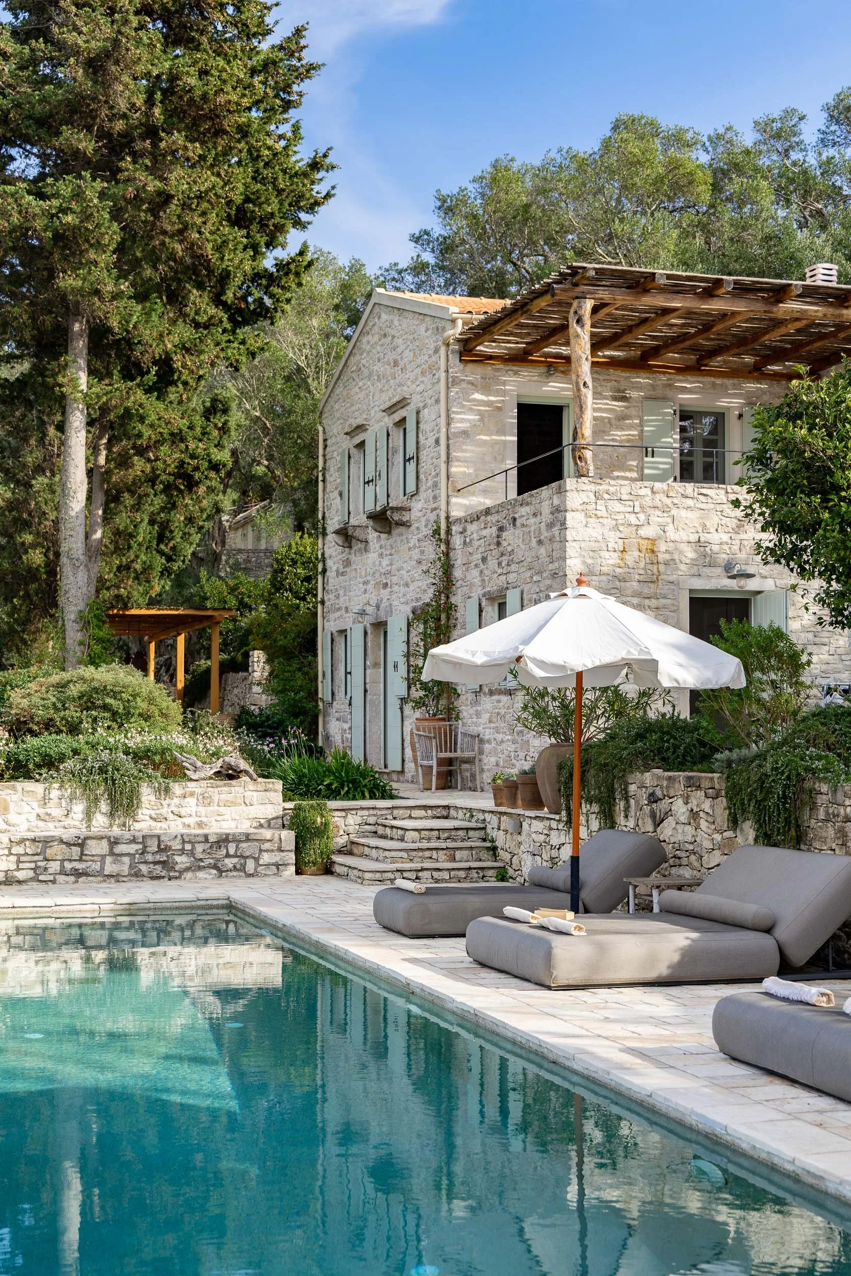 A scenic outdoor view of a stone house with balcony and shutters, surrounded by lush greenery, with a swimming pool in the foreground and poolside loungers with an umbrella.