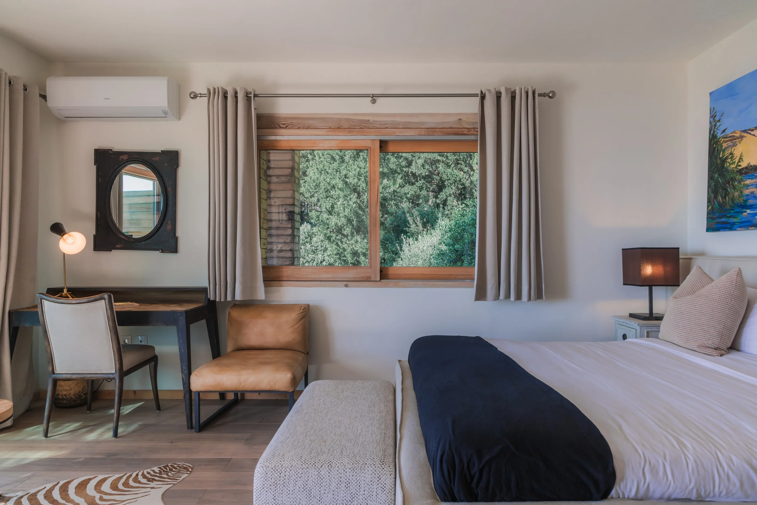 Room with bed, window showing trees, desk, chair, armchair, lamp, and nature painting.