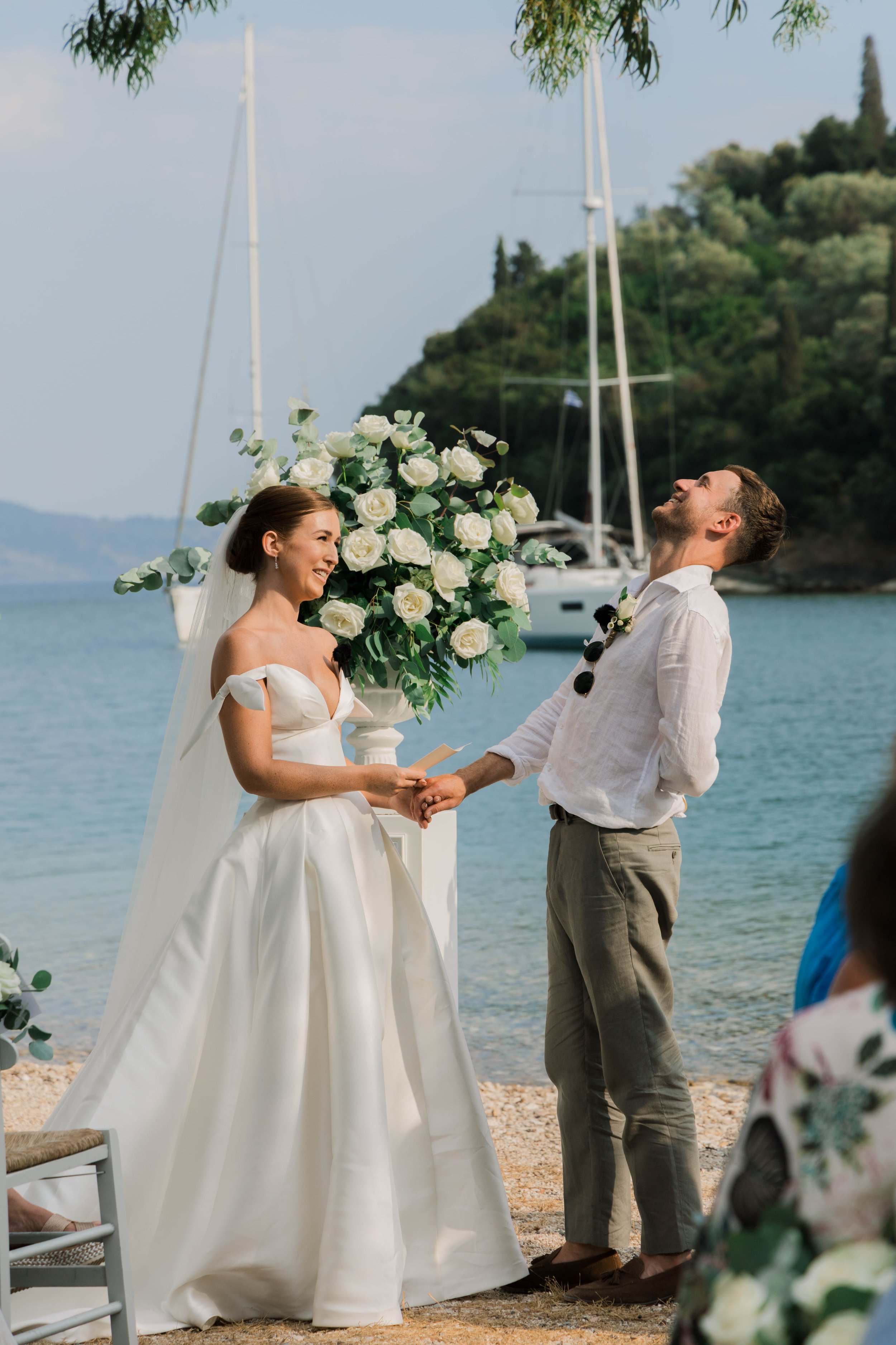 A bride and groom exchange vows at a beach wedding ceremony with sailboats in the background. The bride wears a white wedding gown with bow details on her shoulders, and the groom wears a white shirt and khaki pants. They are holding hands in front o