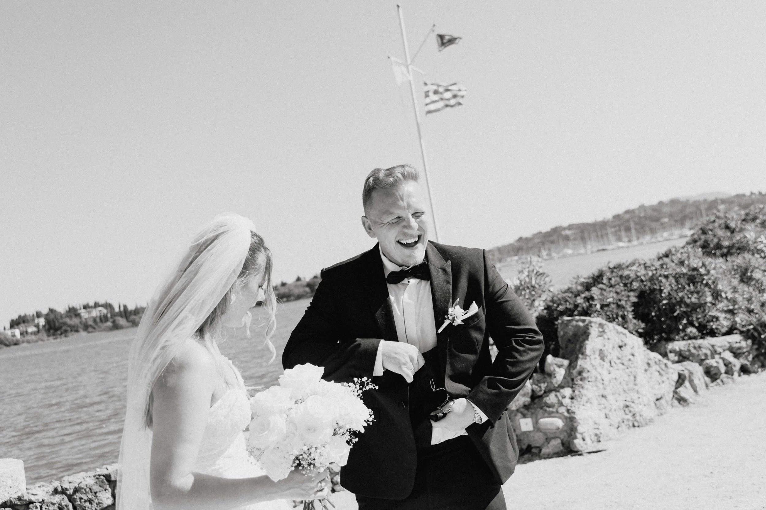 A black and white photo of a bride and groom outdoors by the water, with the groom laughing and the bride holding a bouquet of flowers.