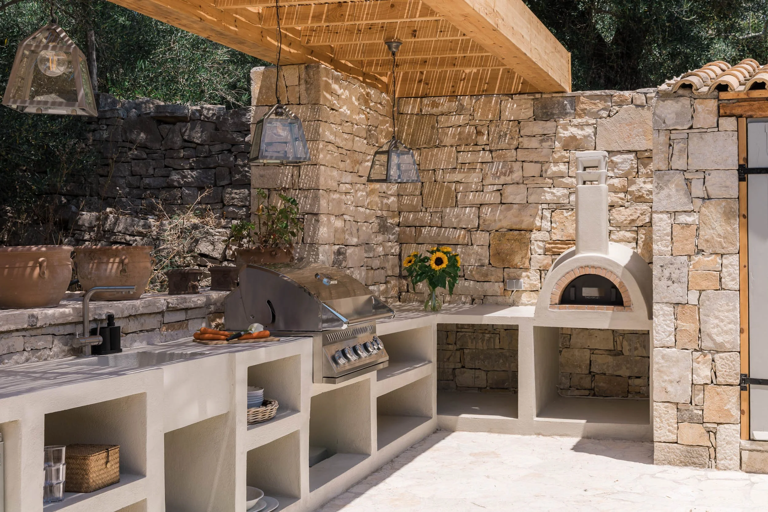 Outdoor kitchen with a stone wall, four hanging lanterns, a built-in grill, a conveyor oven, sunflowers in a vase, and various potted plants.