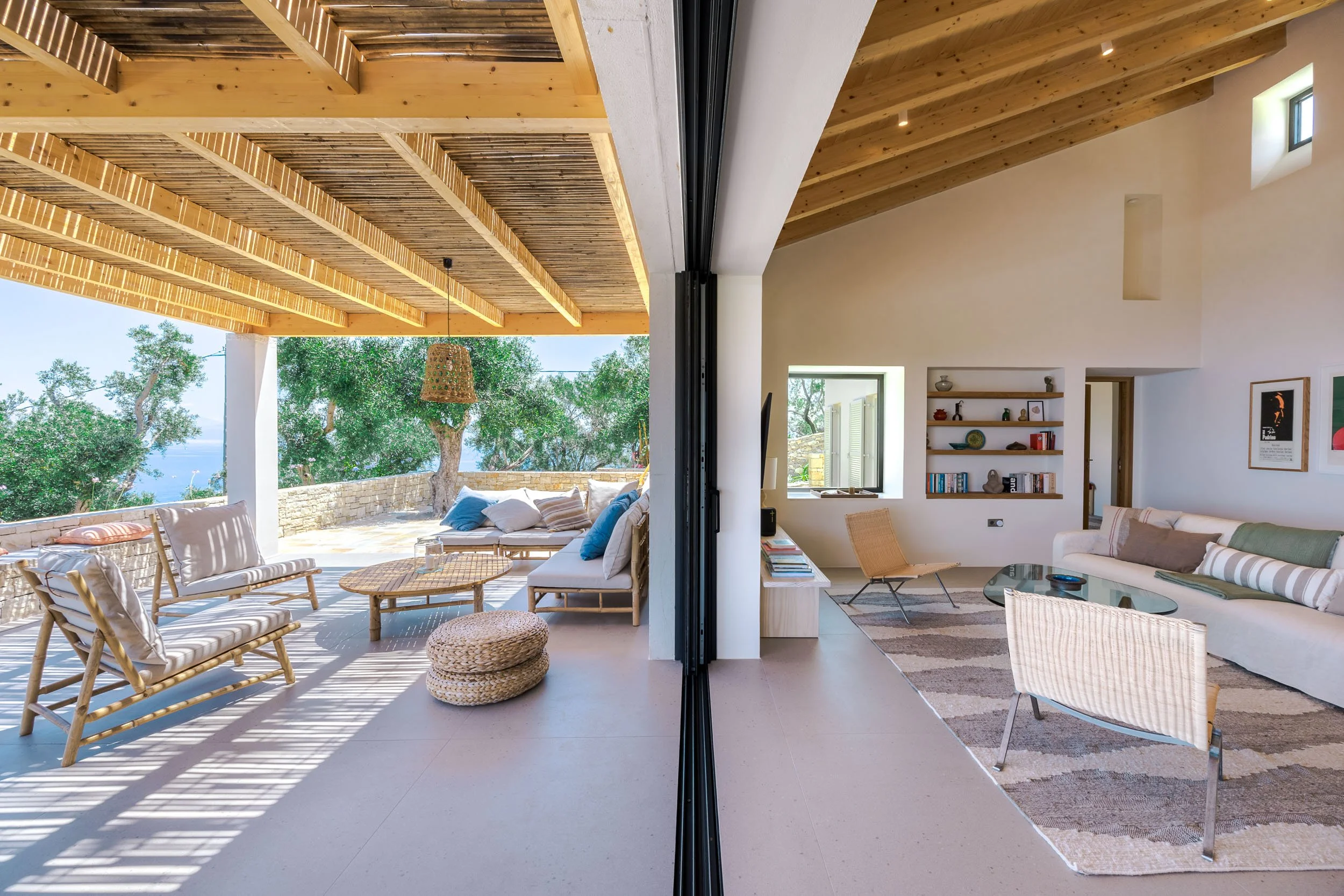 A split view of the interior and exterior of a modern house, with a living room on the right and a covered outdoor patio on the left. The outdoor area features wooden ceiling beams, wicker furniture, and trees with a view of the ocean. The indoor liv