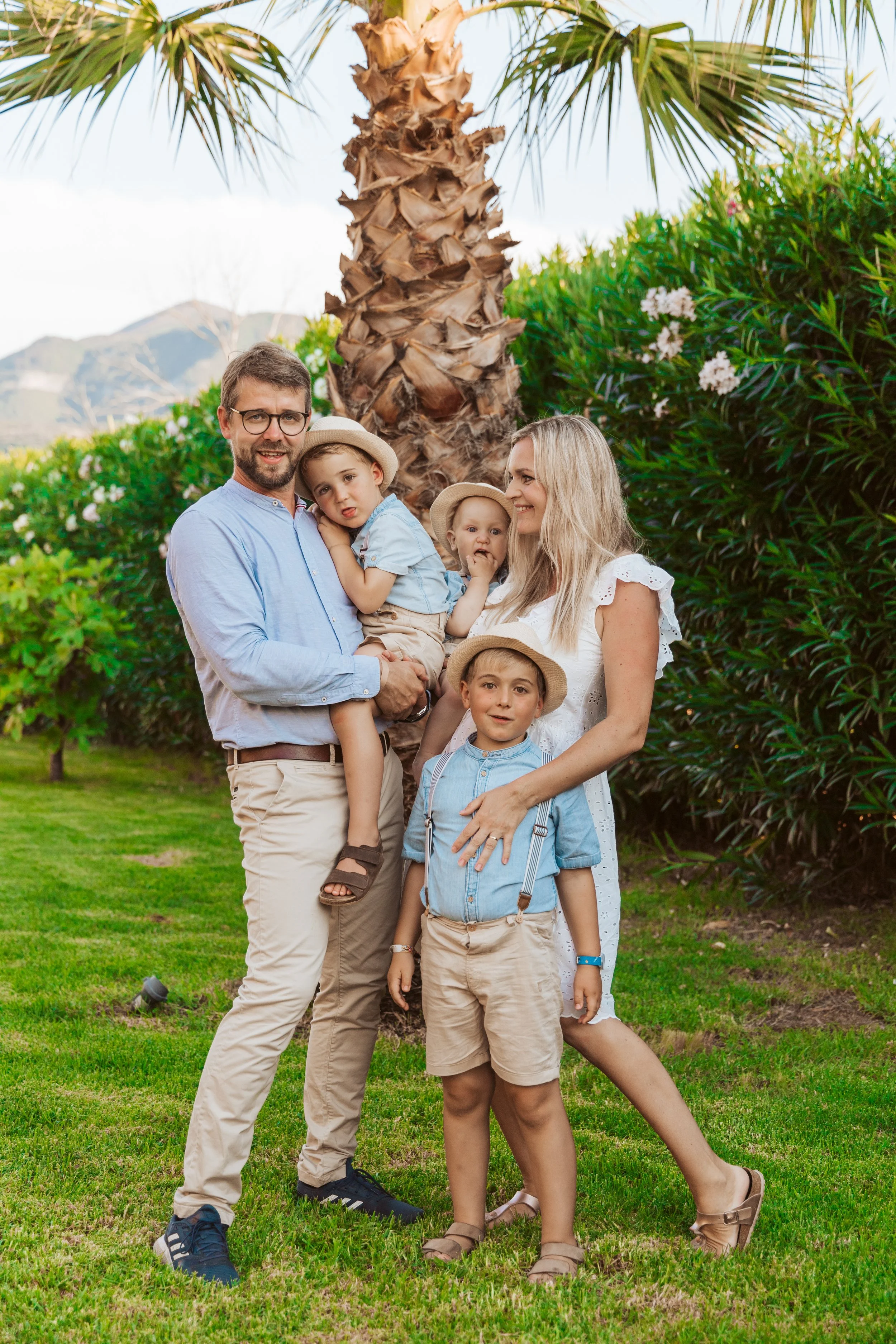 A family of five enjoying time outdoors in a park with green grass and lush bushes, standing in front of a large palm tree. The family members, dressed in light summer clothing, include a man, woman, and three children, some wearing hats.