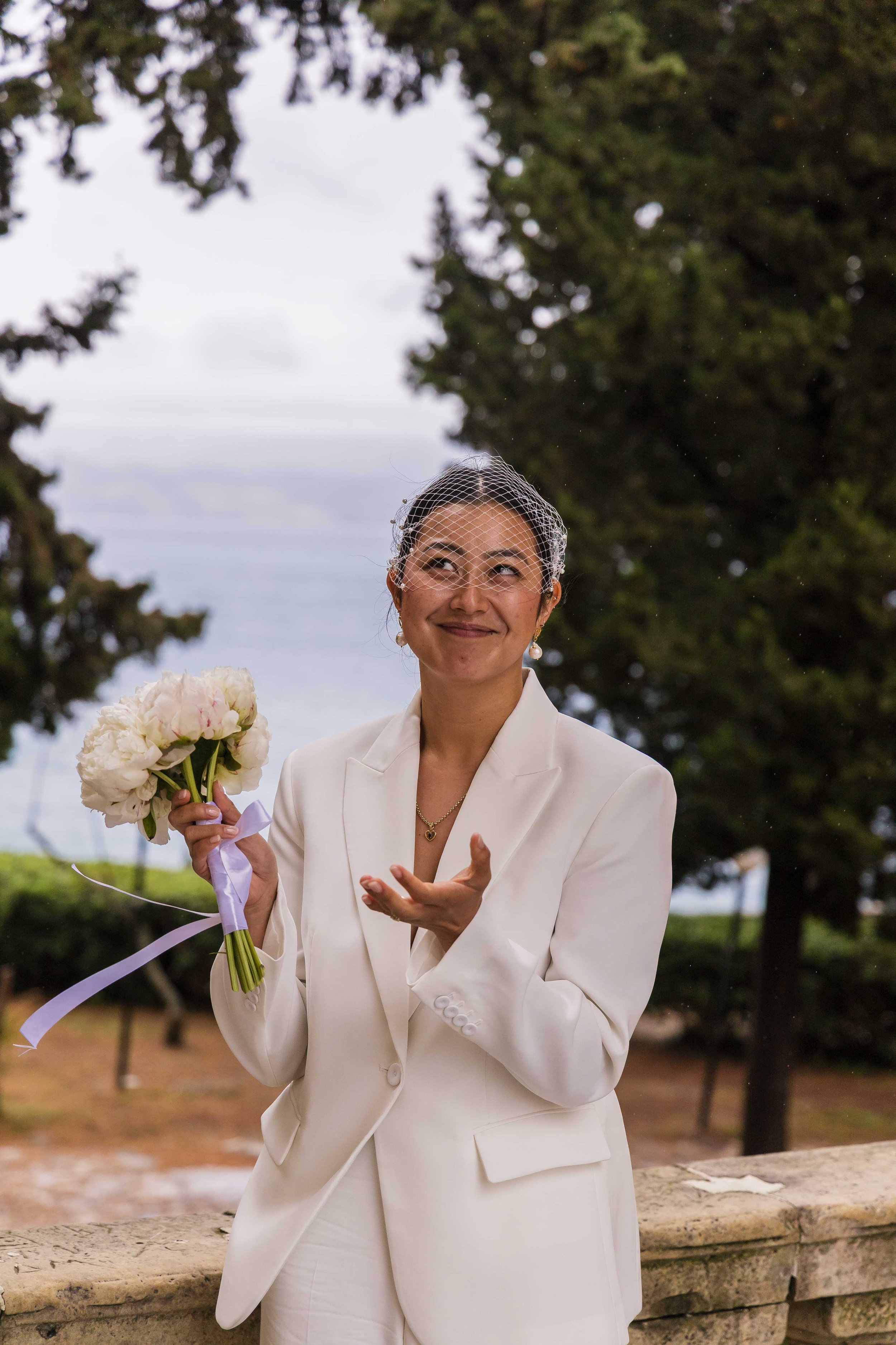 A woman in a white suit with a birdcage veil, holding a flower bouquet, standing outdoors near a stone railing, smiling and looking upward.