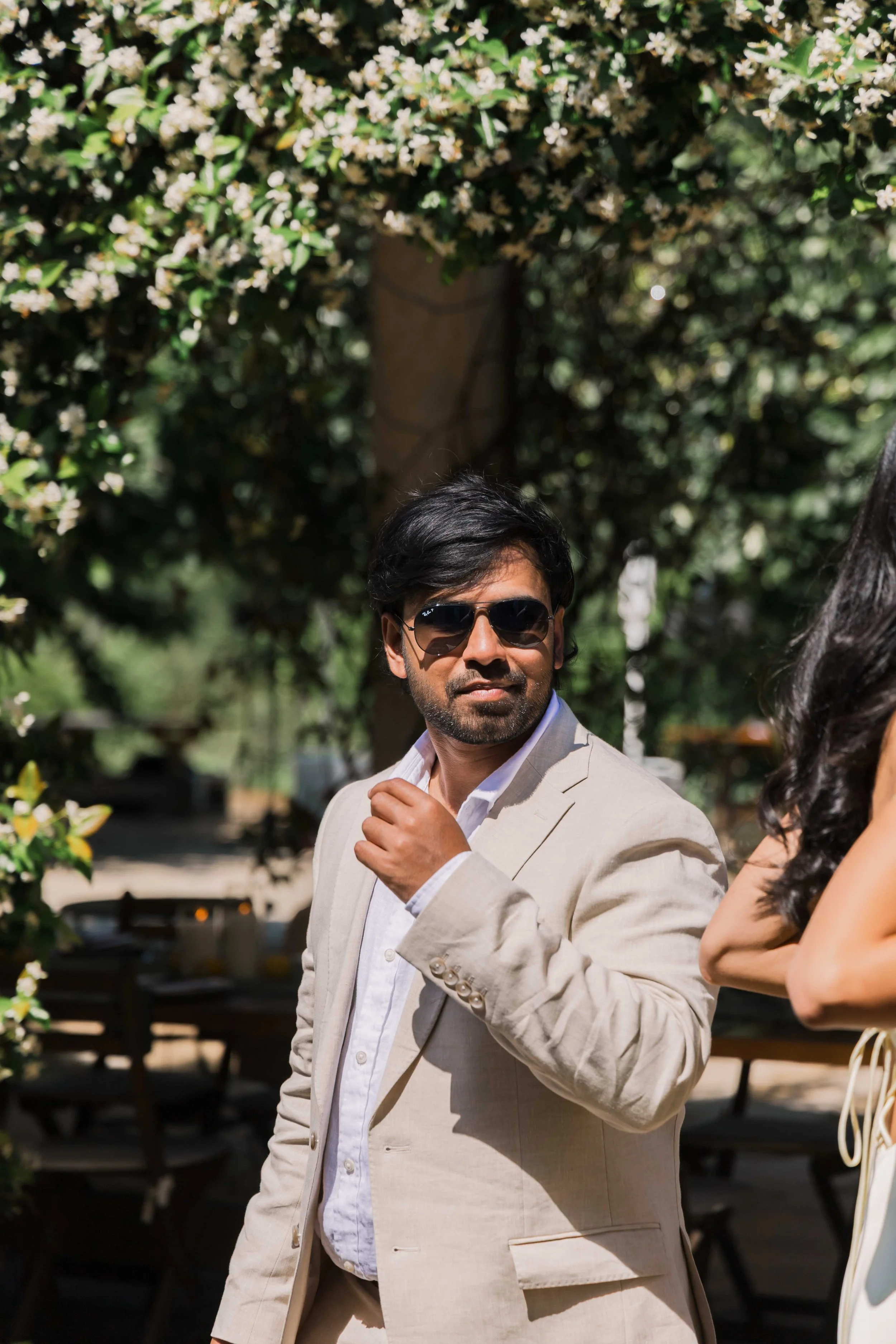 A man in a beige suit and sunglasses standing outdoors amidst greenery, with trees and white flowers overhead.