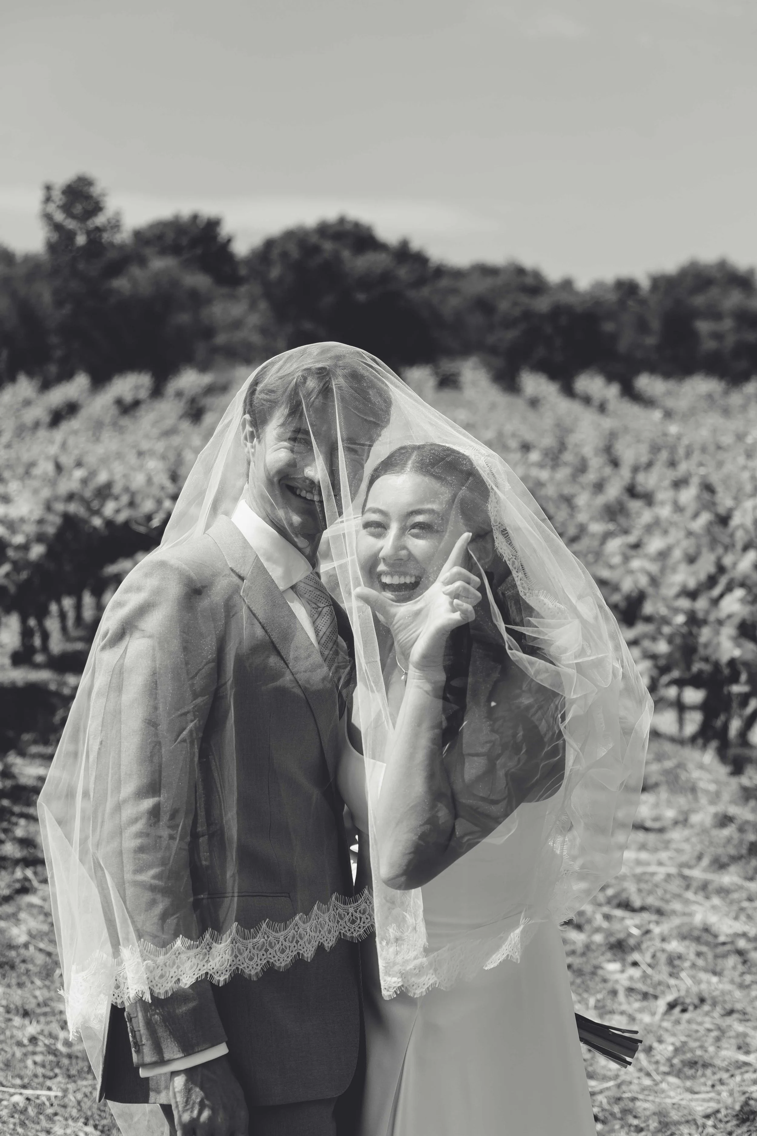 Black and white photo of a newlywed couple smiling and laughing under a wedding veil outdoors, with a vineyard in the background.