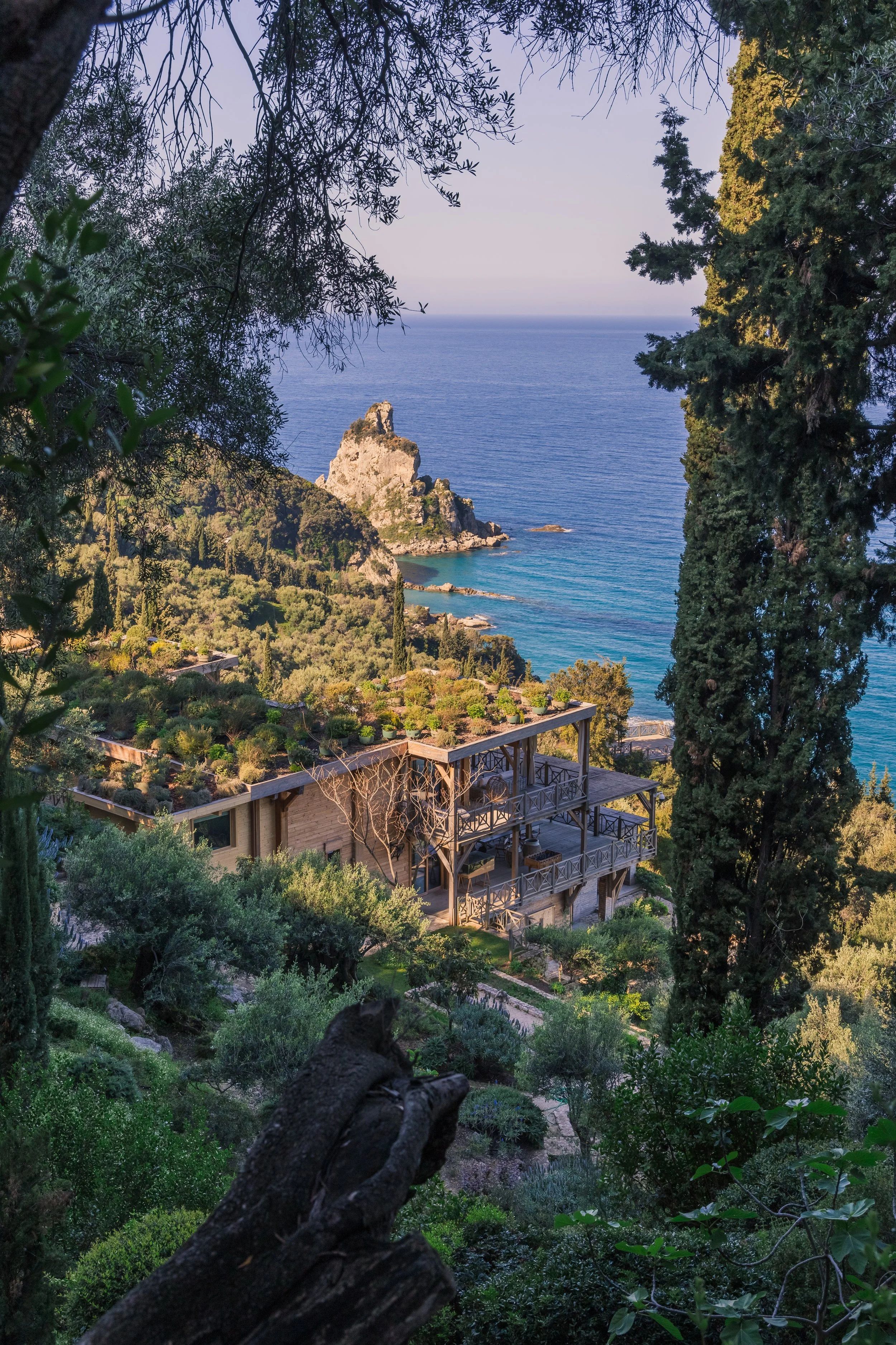 A scenic coastal view of a hillside with a house surrounded by trees, overlooking the sea with a rocky formation in the distance.