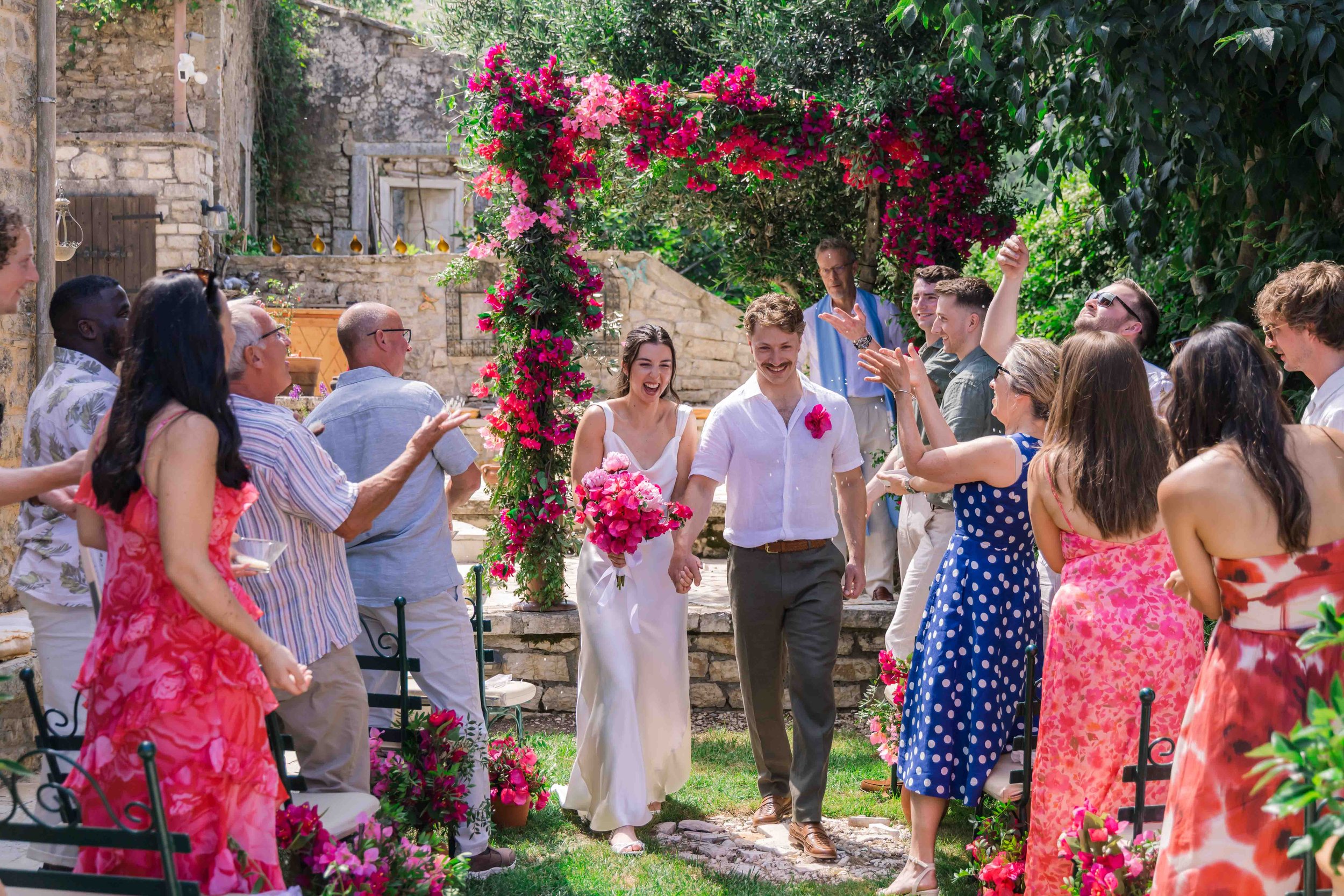 A wedding celebration outdoors with a bride and groom walking hand in hand under a floral archway, surrounded by friends and family clapping and celebrating on a sunny day.