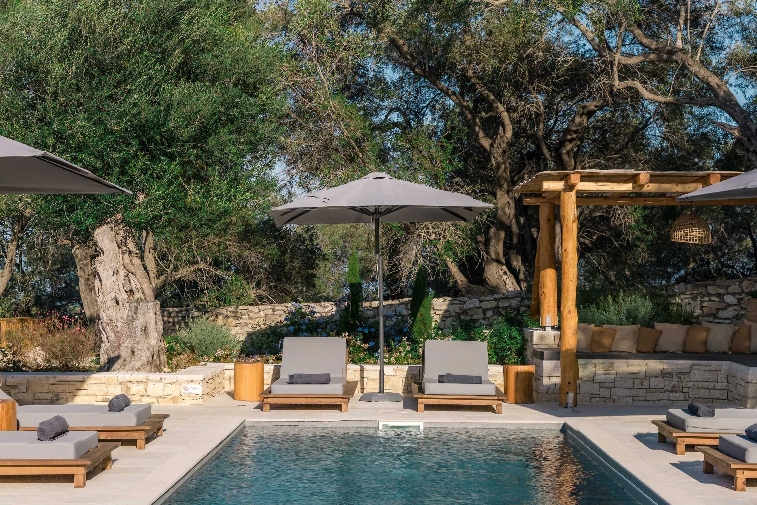 An outdoor pool area with lounge chairs, umbrellas, a stone wall, trees, and a shaded seating area.