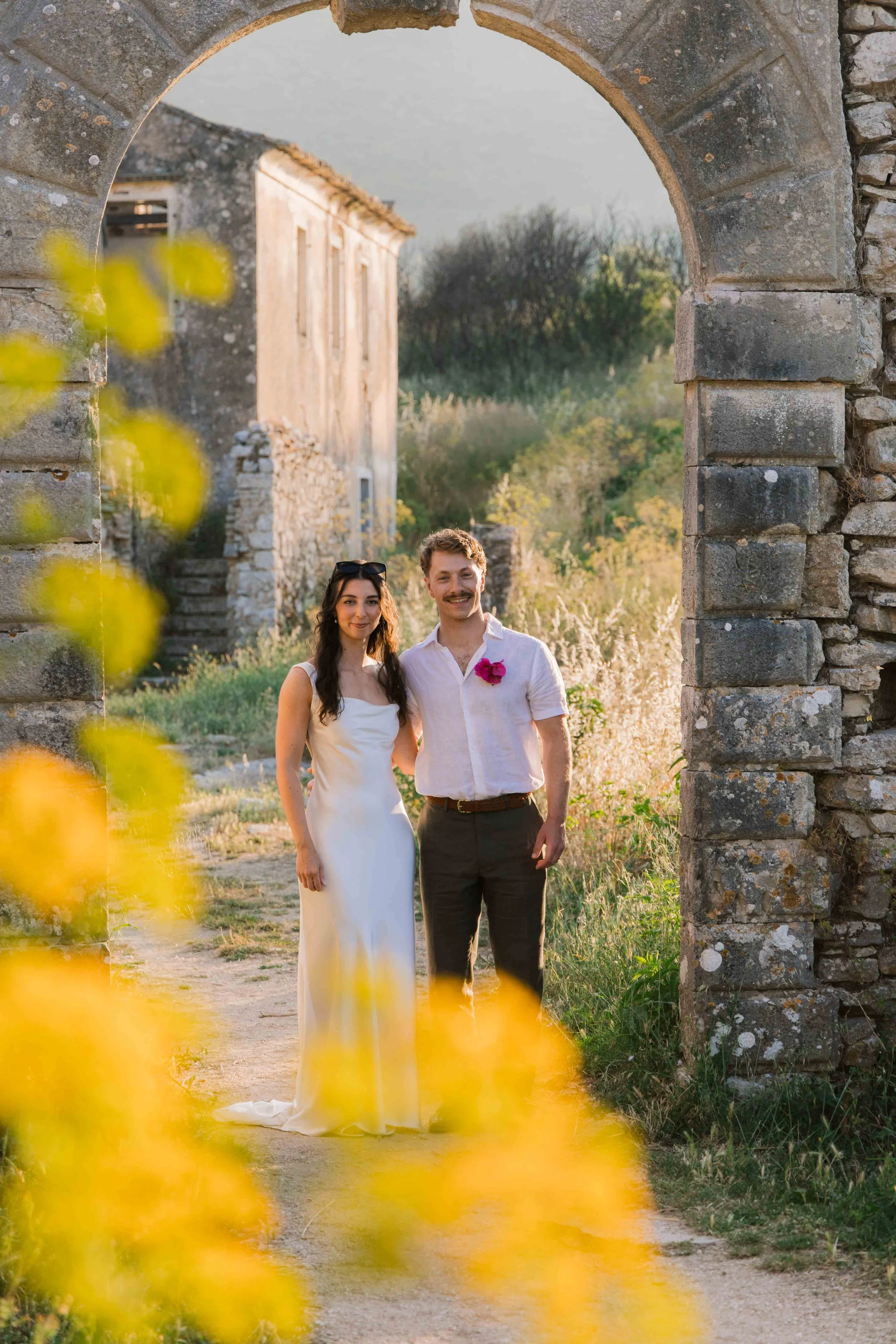 A couple, a woman in a white dress and a man in a white shirt with a pink flower, standing hand-in-hand in front of an ancient stone archway during a sunny day.