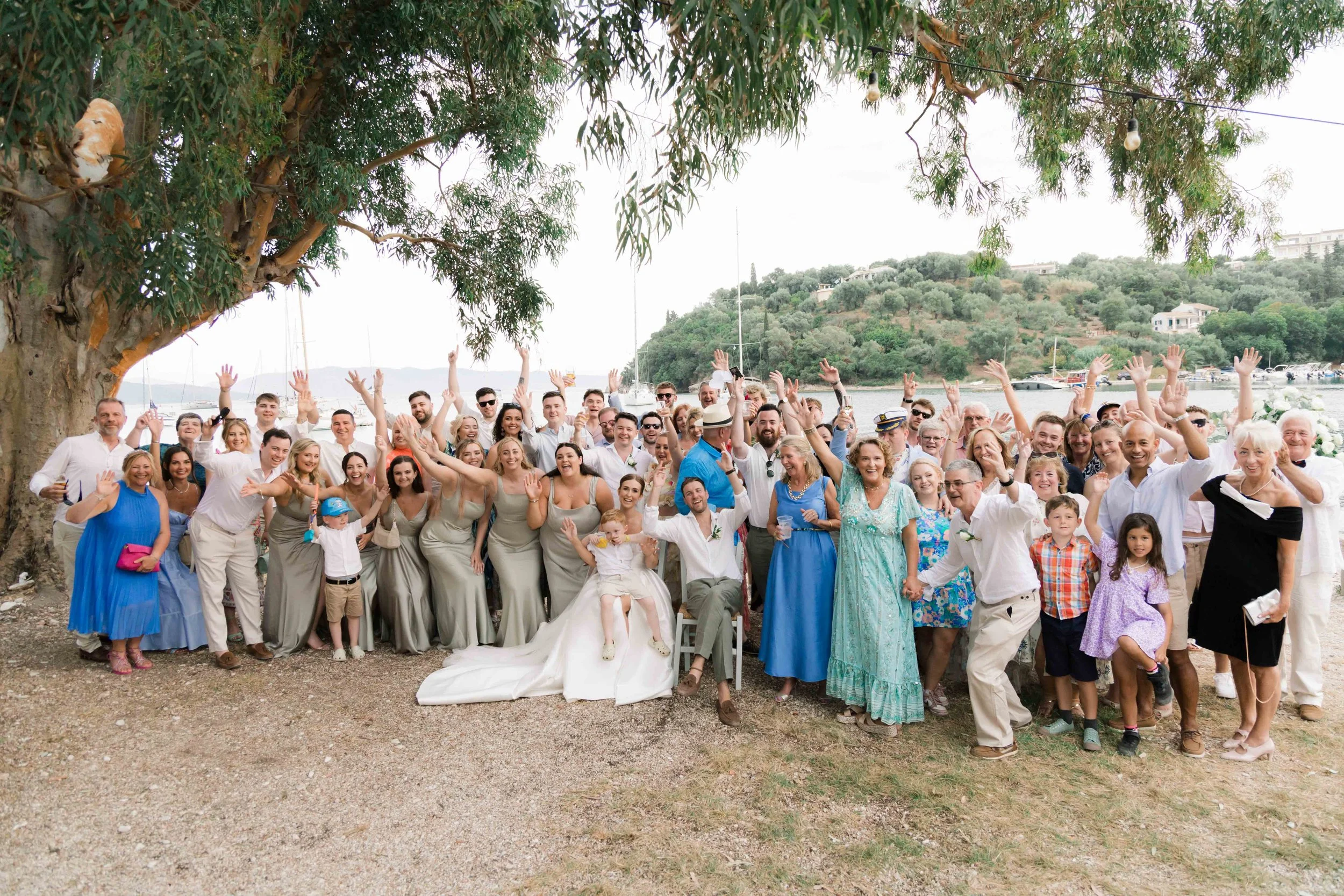 A large group of people celebrating outdoors under a tree near a body of water, with some wearing formal attire, including a bride and groom, and others dressed casually.