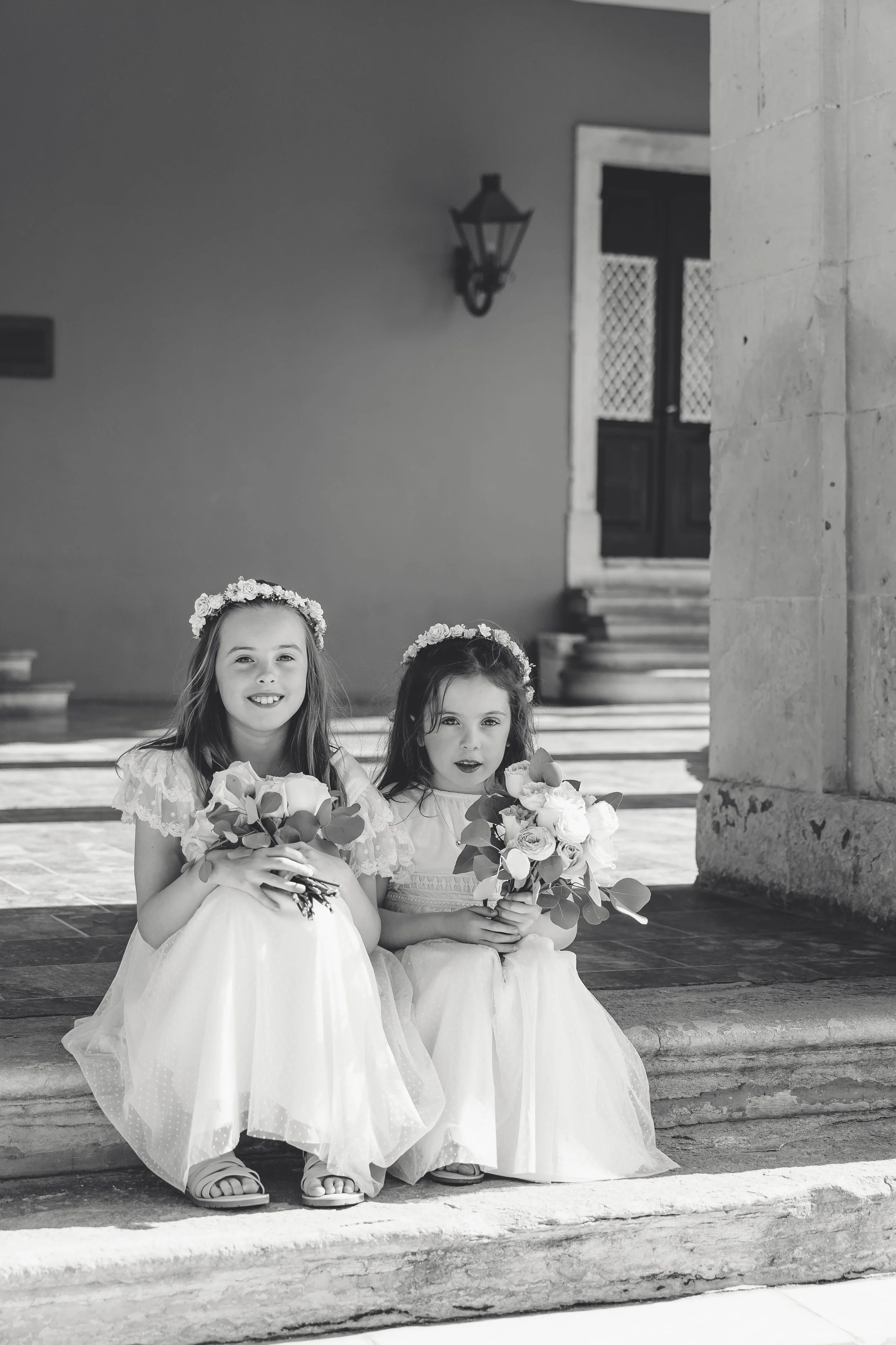 Two young girls in white dresses and floral headbands sitting on steps, holding bouquets, during a wedding or special occasion.