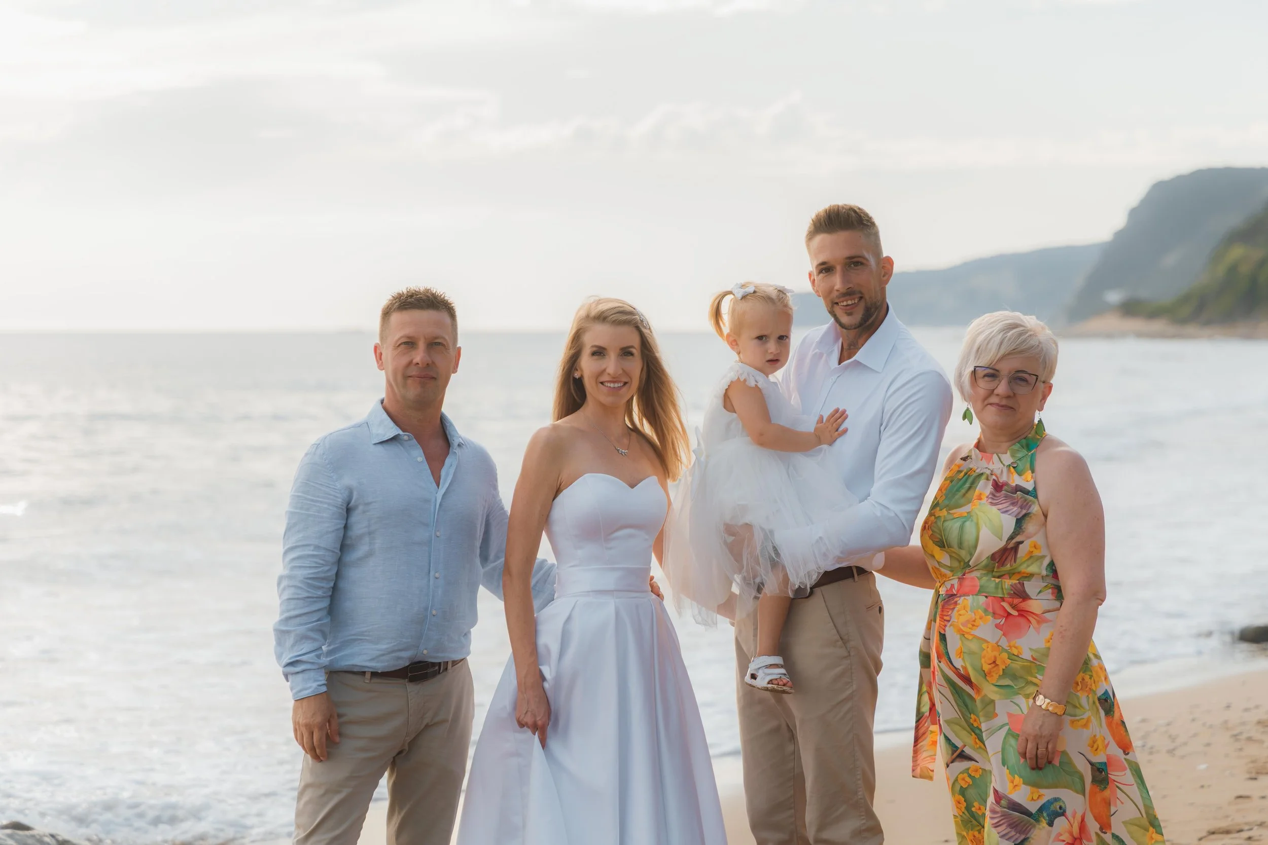 Group of five people, three adults and one young girl, standing on a beach near the ocean, dressed in casual and semi-formal clothing, smiling at the camera.