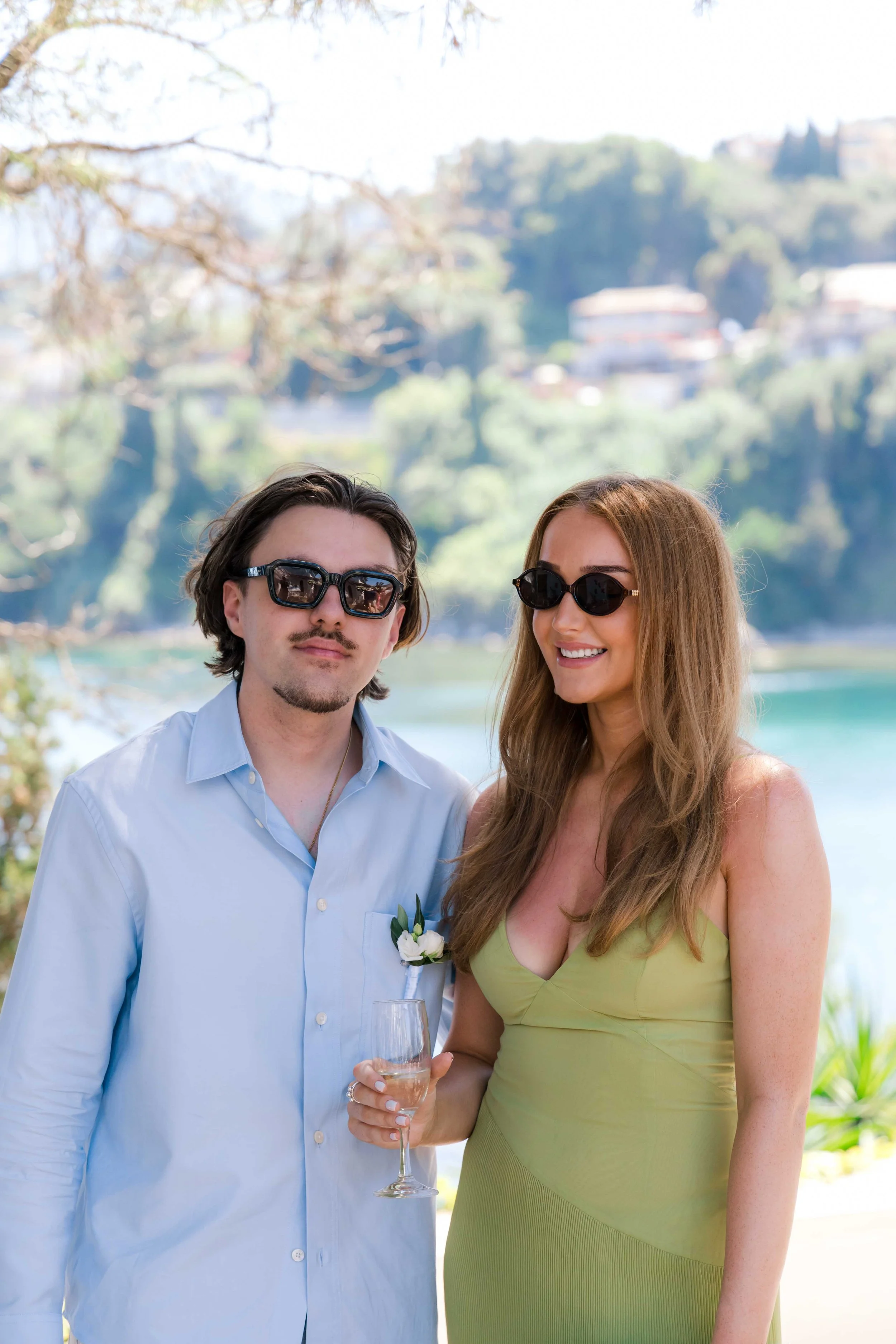 A man and woman standing outdoors by a body of water, with trees and hills in the background. The man is wearing sunglasses and a light blue shirt with a boutonnière. The woman is wearing sunglasses and a light green dress, holding a glass of champag