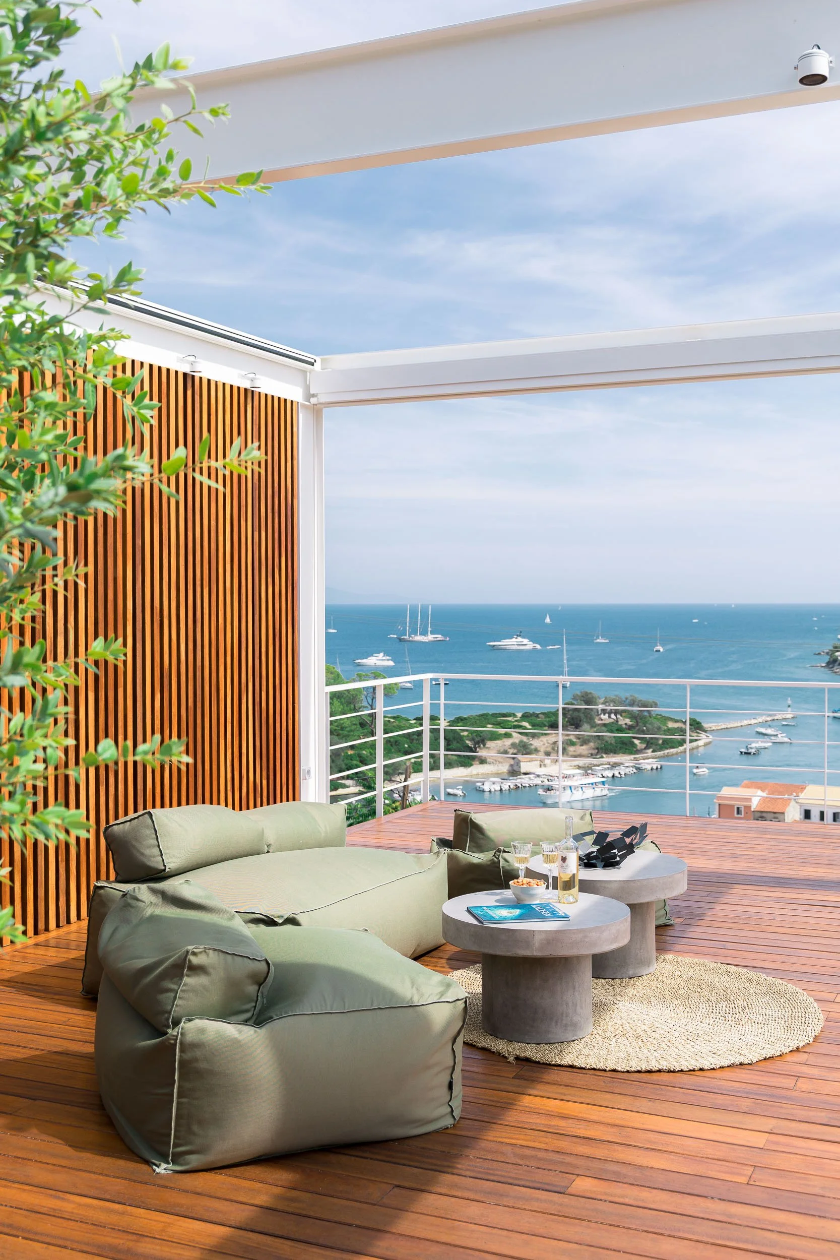 A spacious outdoor balcony with wooden flooring, green cushioned seating, two round concrete tables with drinks and snacks, overlooking a scenic view of the ocean with boats and sailboats, surrounded by lush greenery and a clear sky.