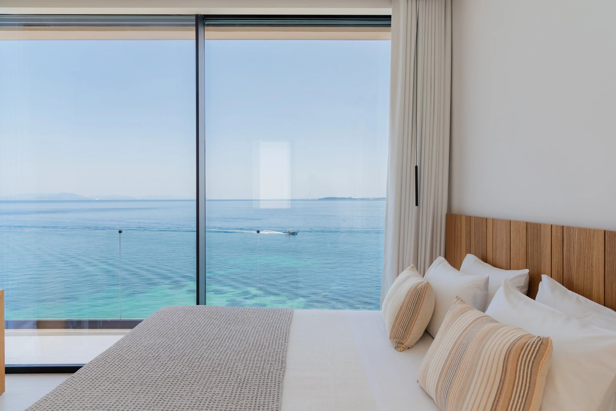 Modern bedroom with a large window view of the ocean and a boat passing by, featuring a wooden headboard, white bedding, and beige striped pillows.
