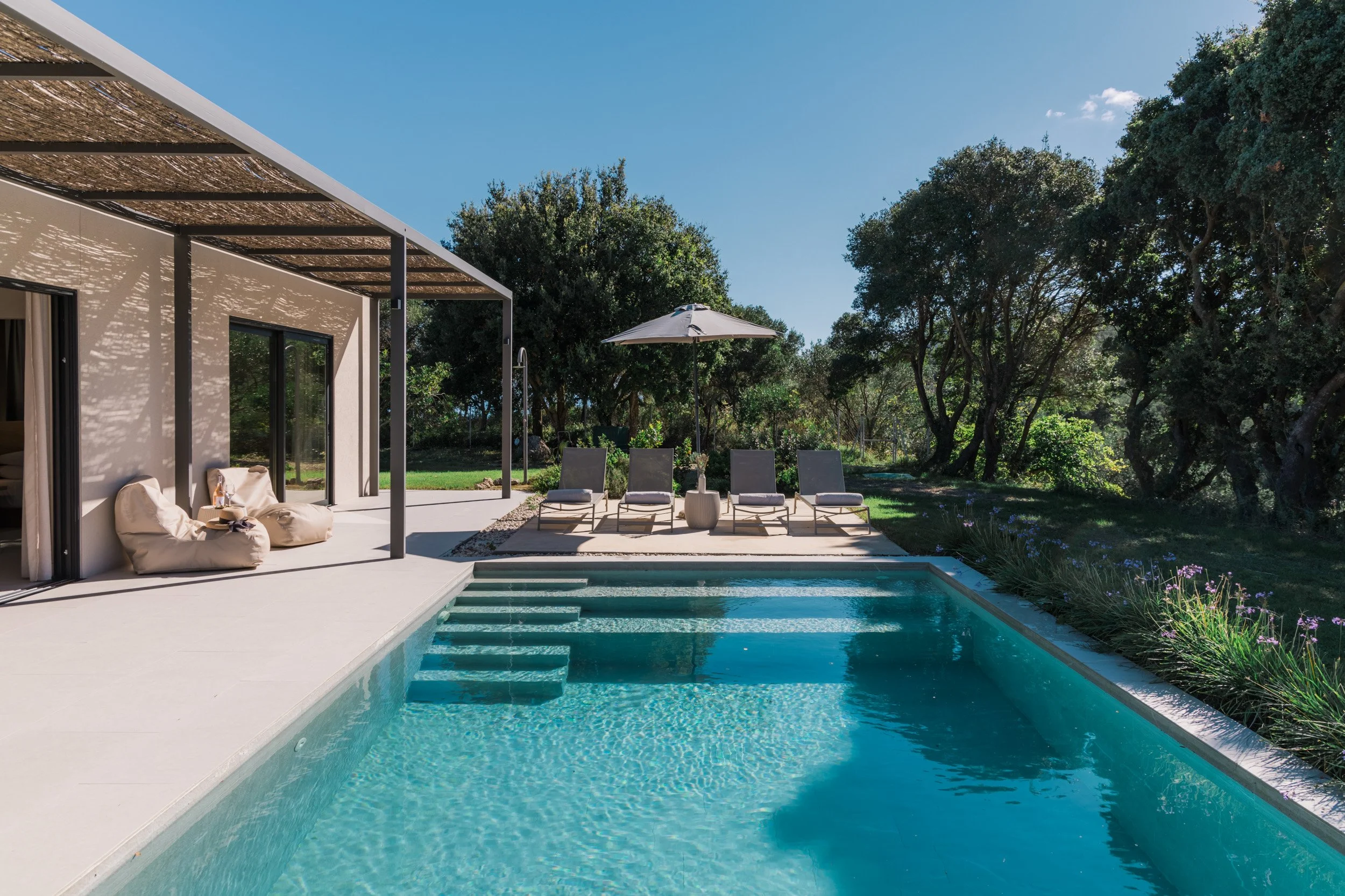 Modern backyard pool area with lounge chairs, an umbrella, and a shaded patio space surrounded by tall trees.
