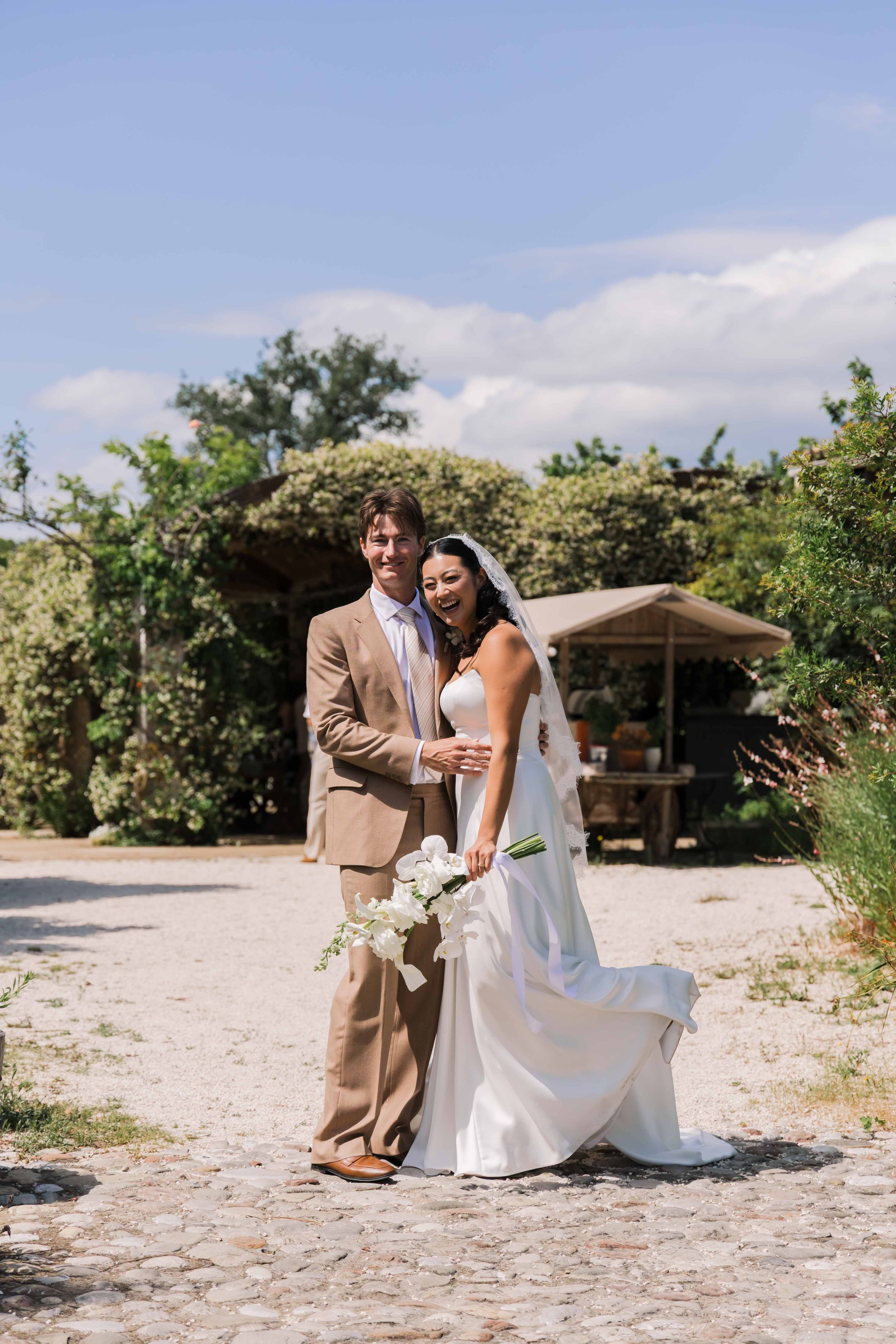 A newlywed couple standing on a cobblestone pathway outdoors, smiling and embracing. The bride is holding a white bouquet and wearing a white wedding gown with a veil, while the groom is dressed in a beige suit. There are green trees and bushes, a sm