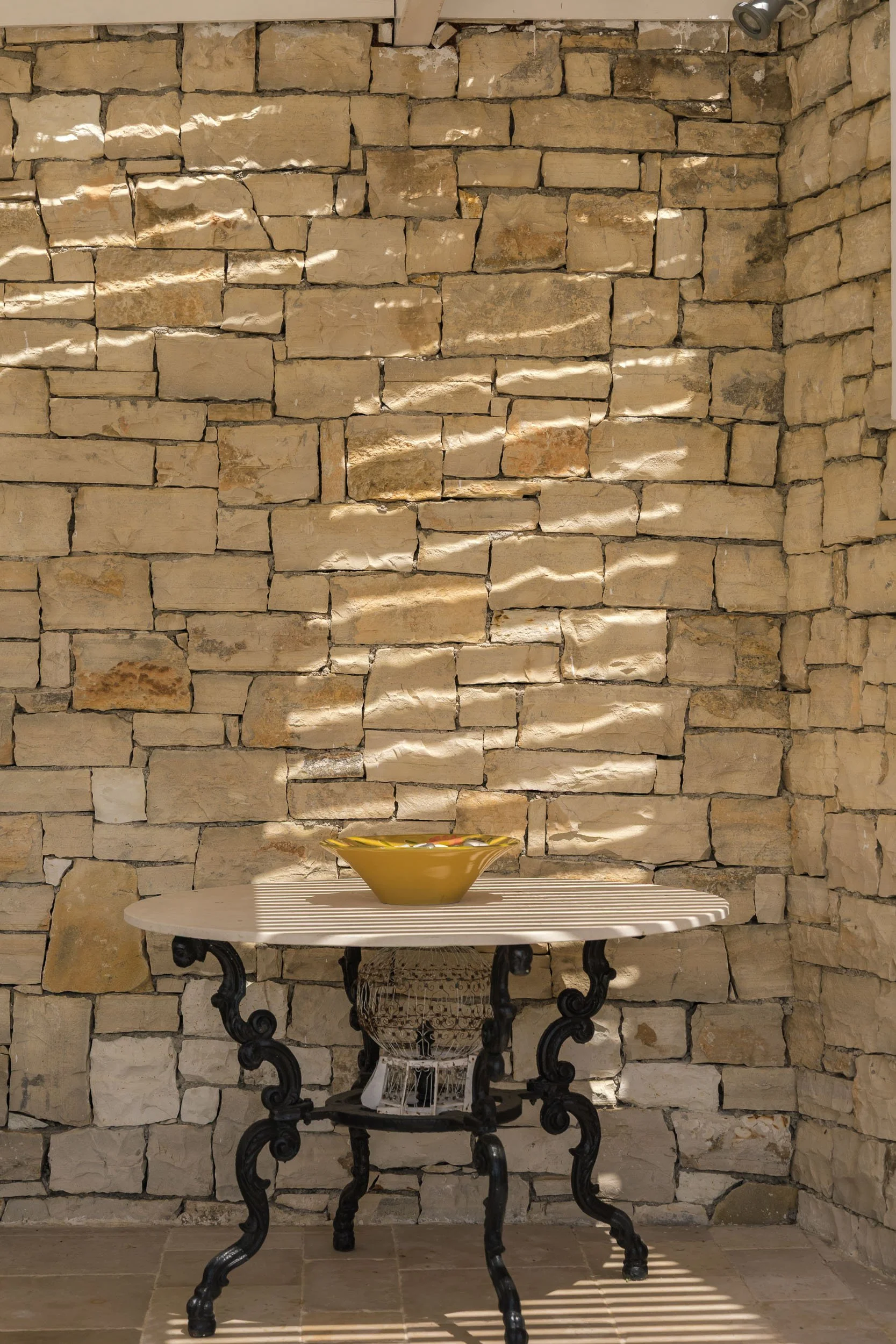 A small round table with a white slatted top and ornate black legs, set against a stone wall. A yellow bowl is placed in the center of the table, with sunlight casting shadows through a striped pattern.