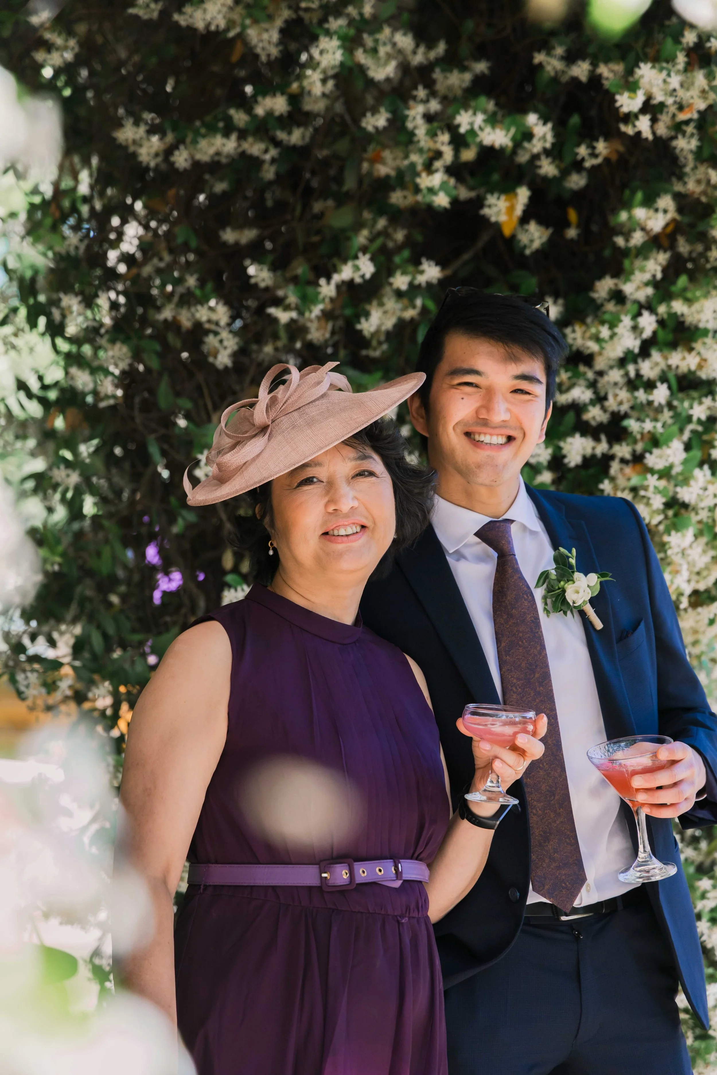 A woman and a young man smile while standing in front of a flowering bush, holding pink cocktails in coupe glasses. The woman wears a purple dress and a large, pink hat, and the man wears a dark suit with a boutonniere.