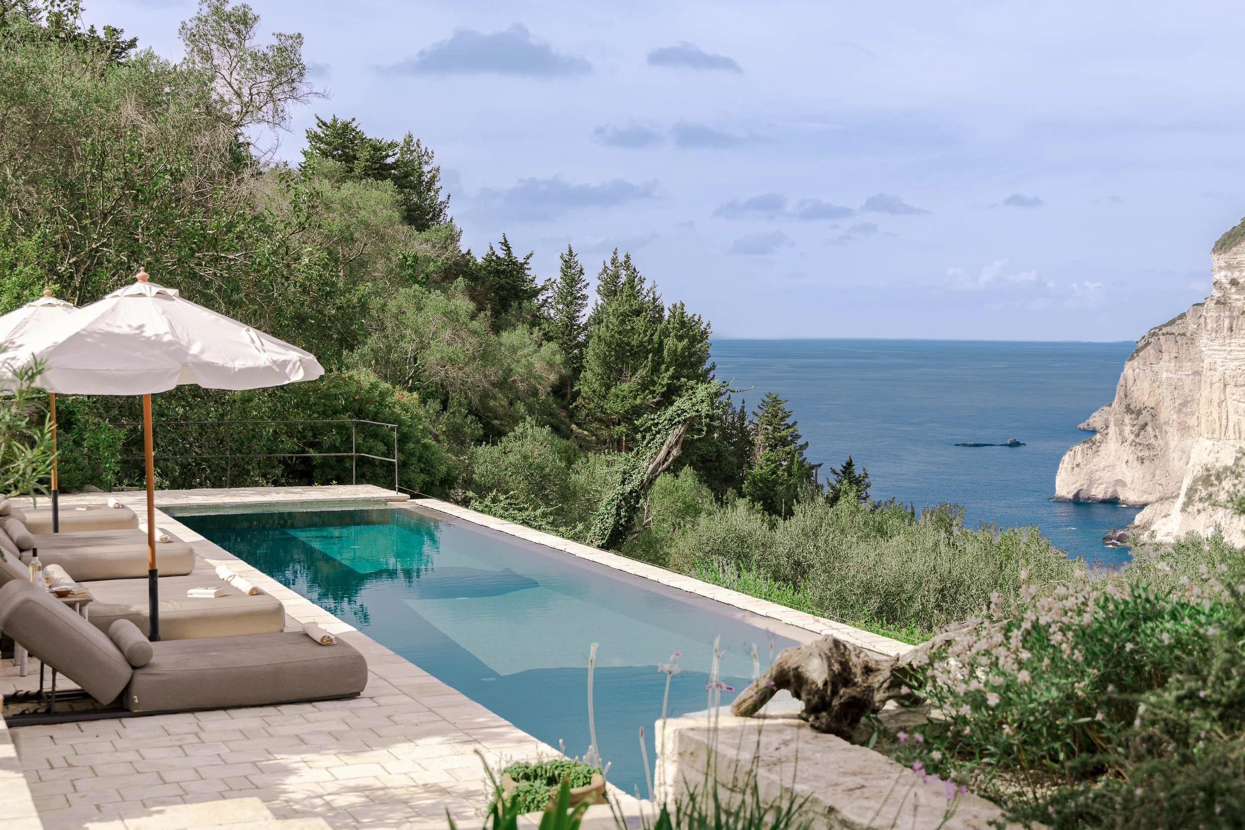 A luxurious outdoor pool area with lounge chairs and umbrellas overlooking the ocean, surrounded by lush greenery and cliffs.