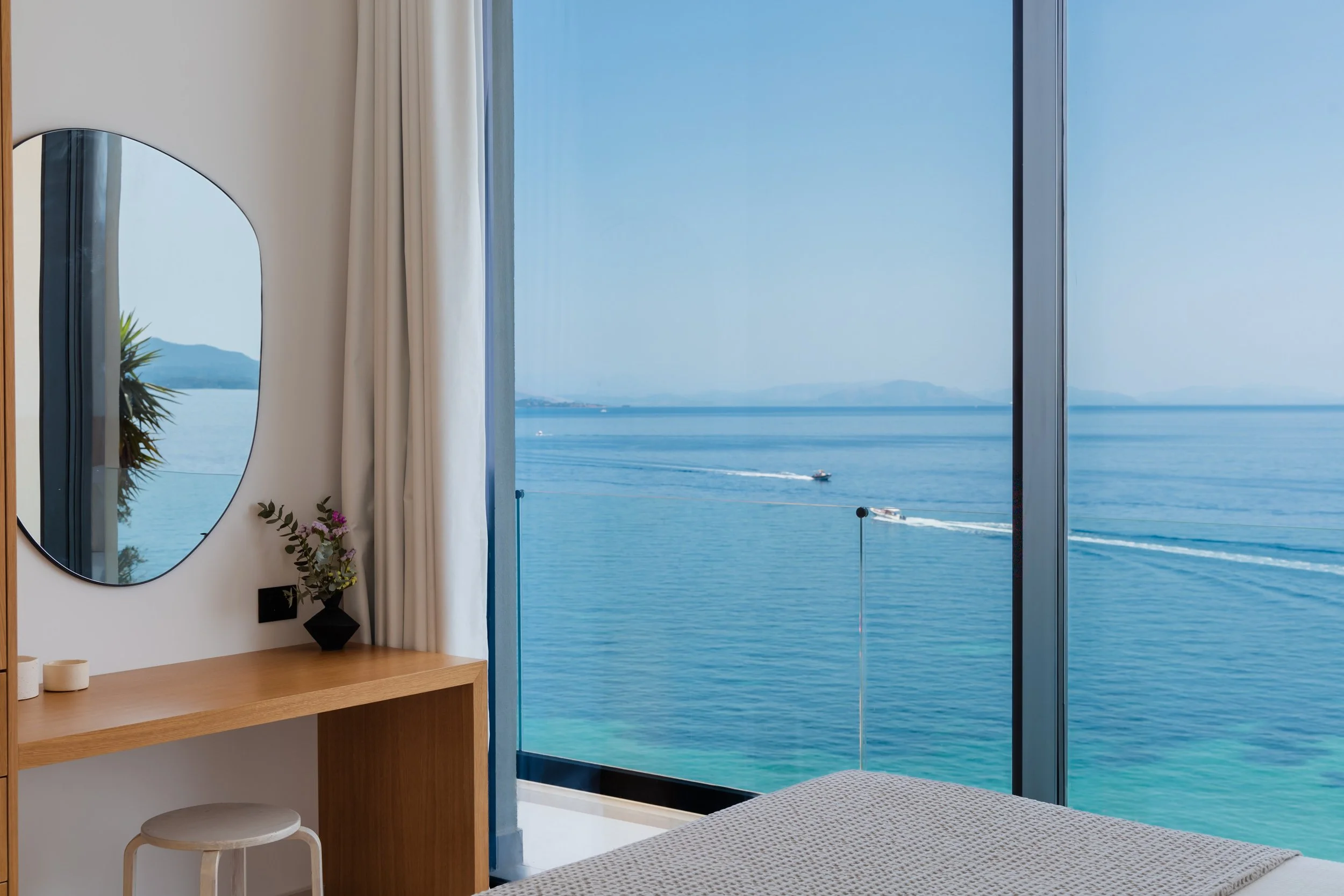 Room with a large glass window overlooking the ocean, with boats sailing on the water, a white curtain, a mirror, a wooden desk with a potted plant, and part of a bed.