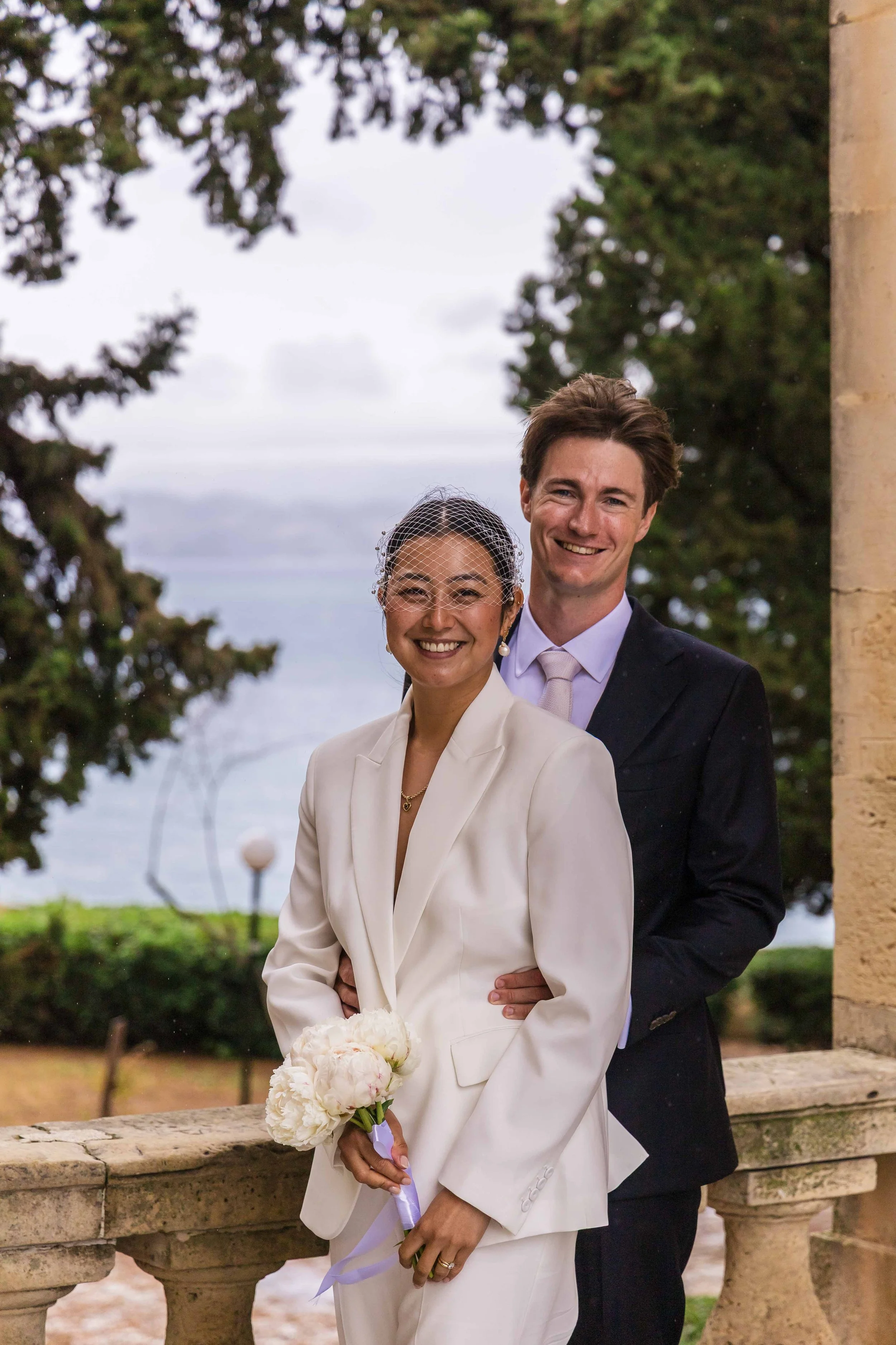 A couple in wedding attire standing outdoors by a stone balustrade, smiling at the camera. The woman is in a white suit and holding a bouquet of white flowers. The man is in a dark suit and a light-colored tie. There are trees and a cloudy sky in the