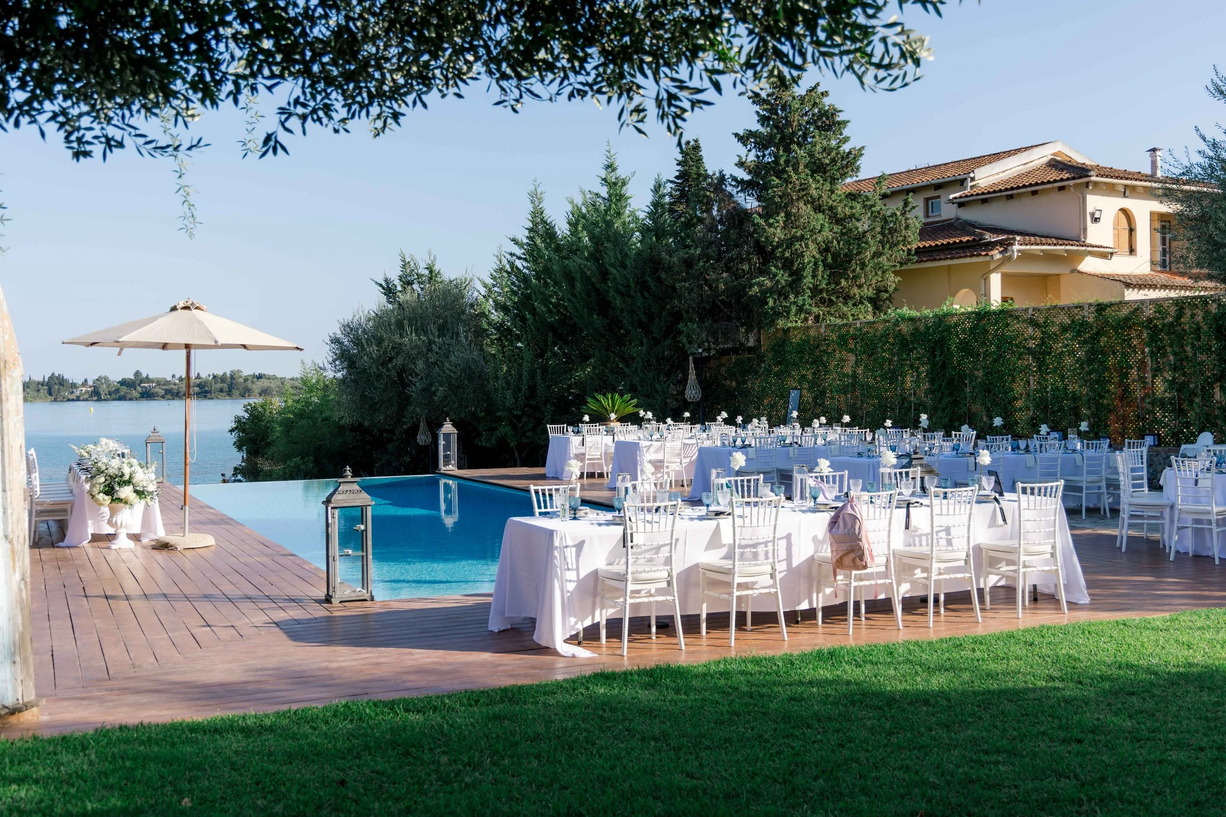 Outdoor wedding reception near a swimming pool with white tables and chairs, floral decorations, an umbrella, and water in the background.