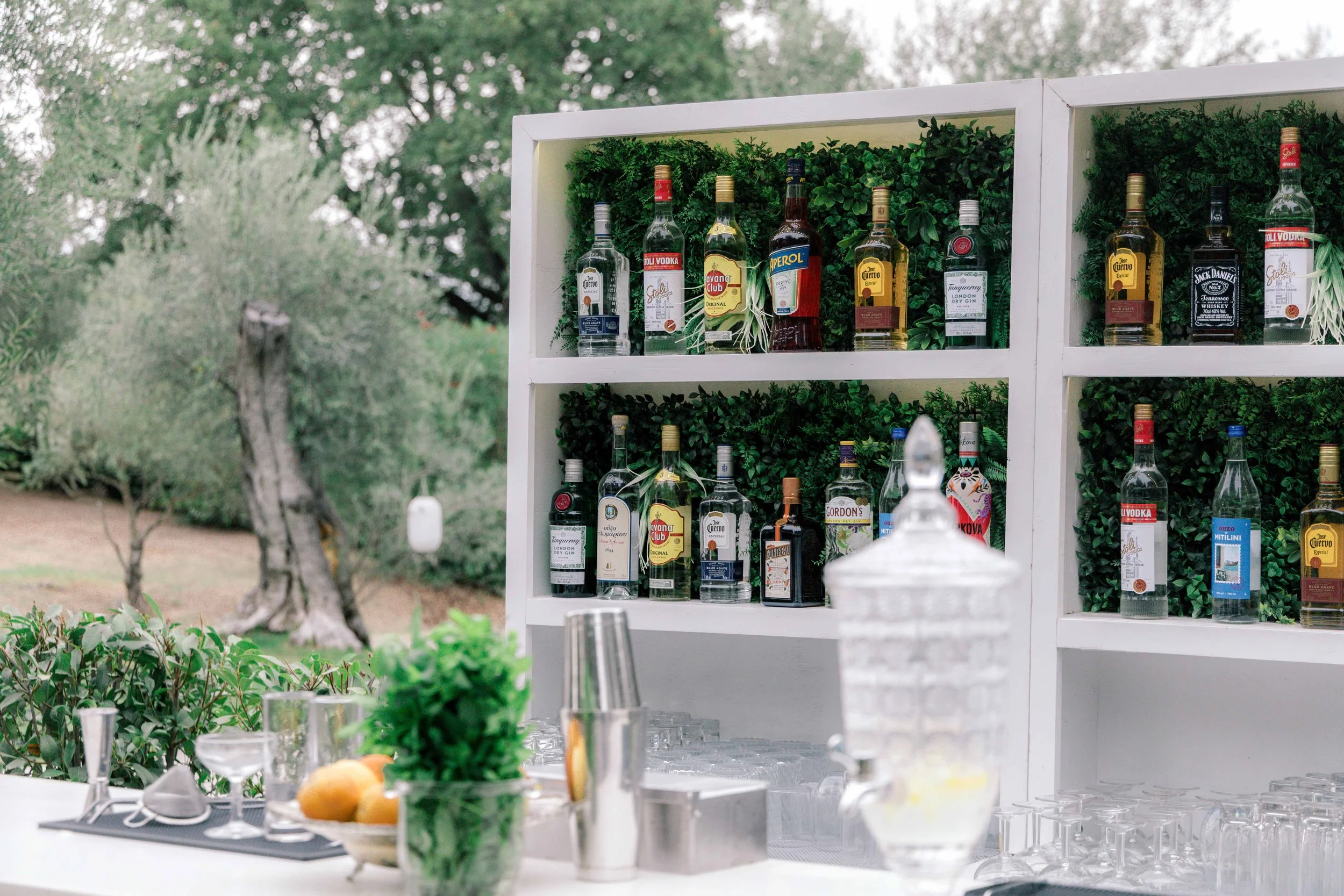 Outdoor bar setup with bottles of alcohol on white shelves against greenery, a pitcher of water, glasses, and fresh fruits and herbs on the bar counter.