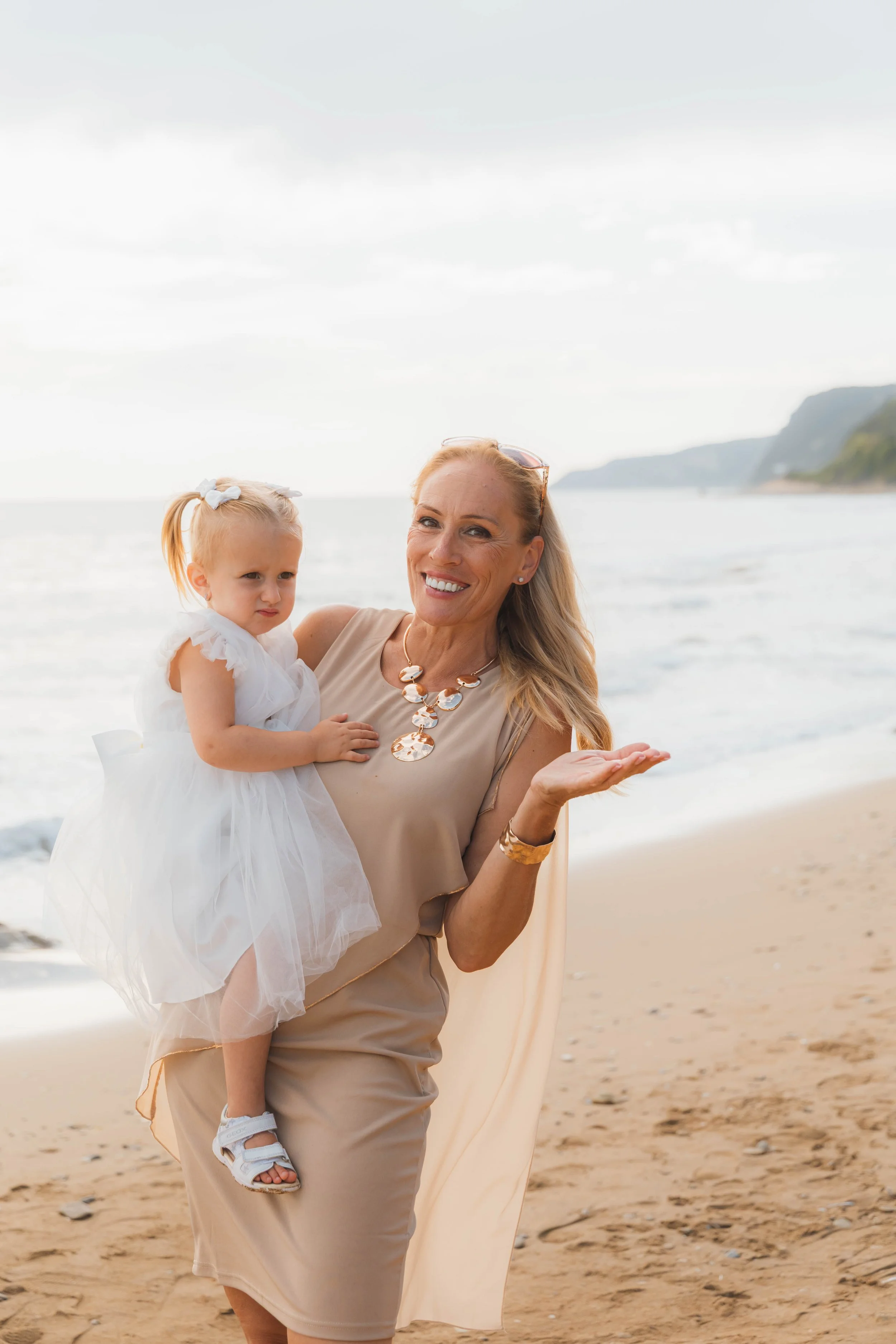A woman holding a young girl on a beach, both smiling, with ocean and cliffs in the background.