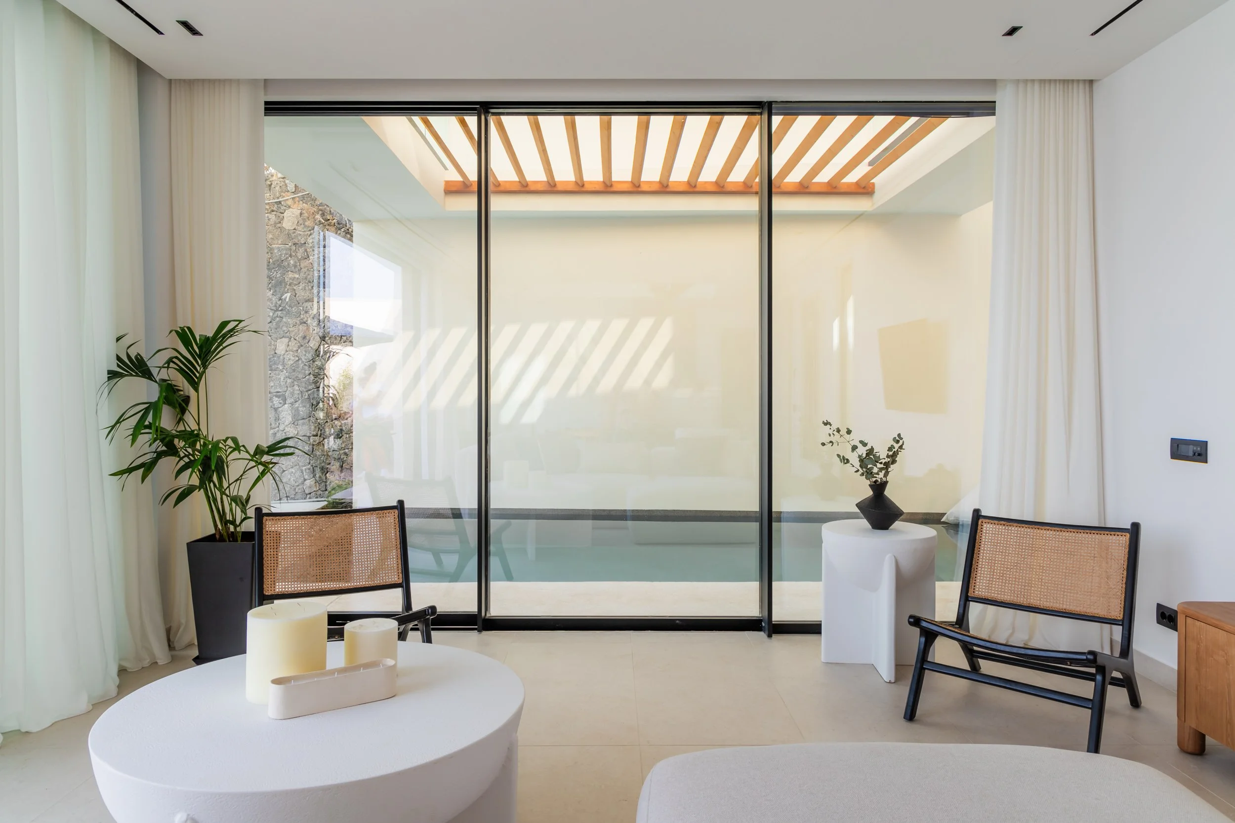 Modern living room with glass sliding door leading to an outdoor patio, decorated with potted plants, a white coffee table with candles, and minimalist furniture.