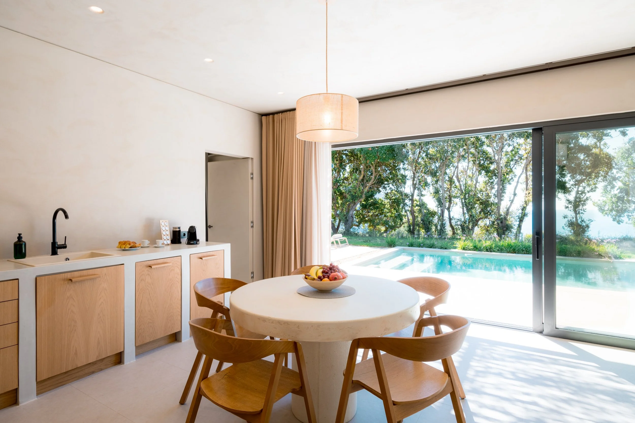 Modern dining area with a white round table, four wooden chairs, a bowl of fruit, a kitchenette with a sink, and large sliding glass doors opening to an outdoor pool and lush greenery.