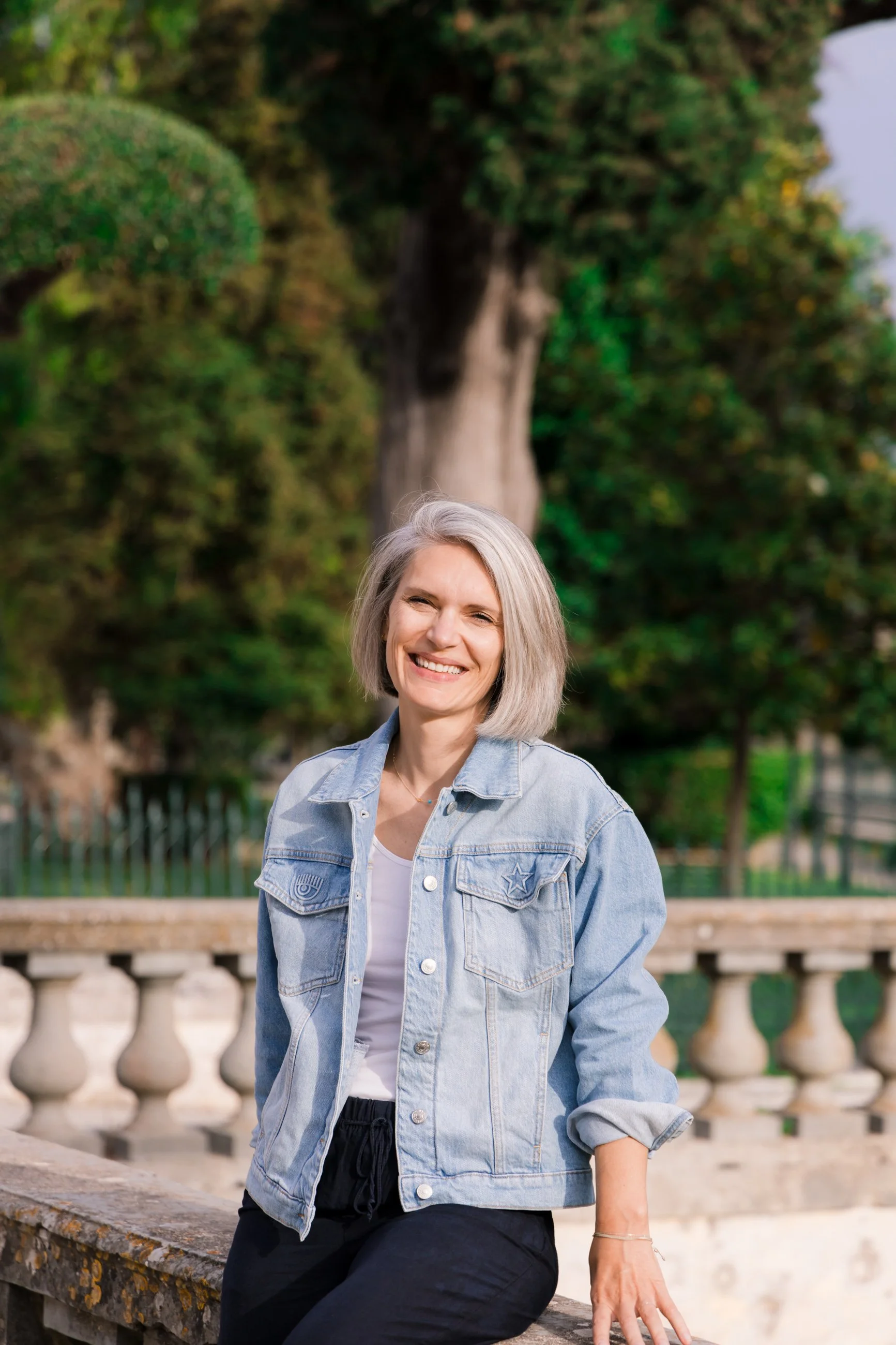 A woman with gray hair smiling outdoors, wearing a light denim jacket and black pants, standing near a stone railing with lush green trees in the background.