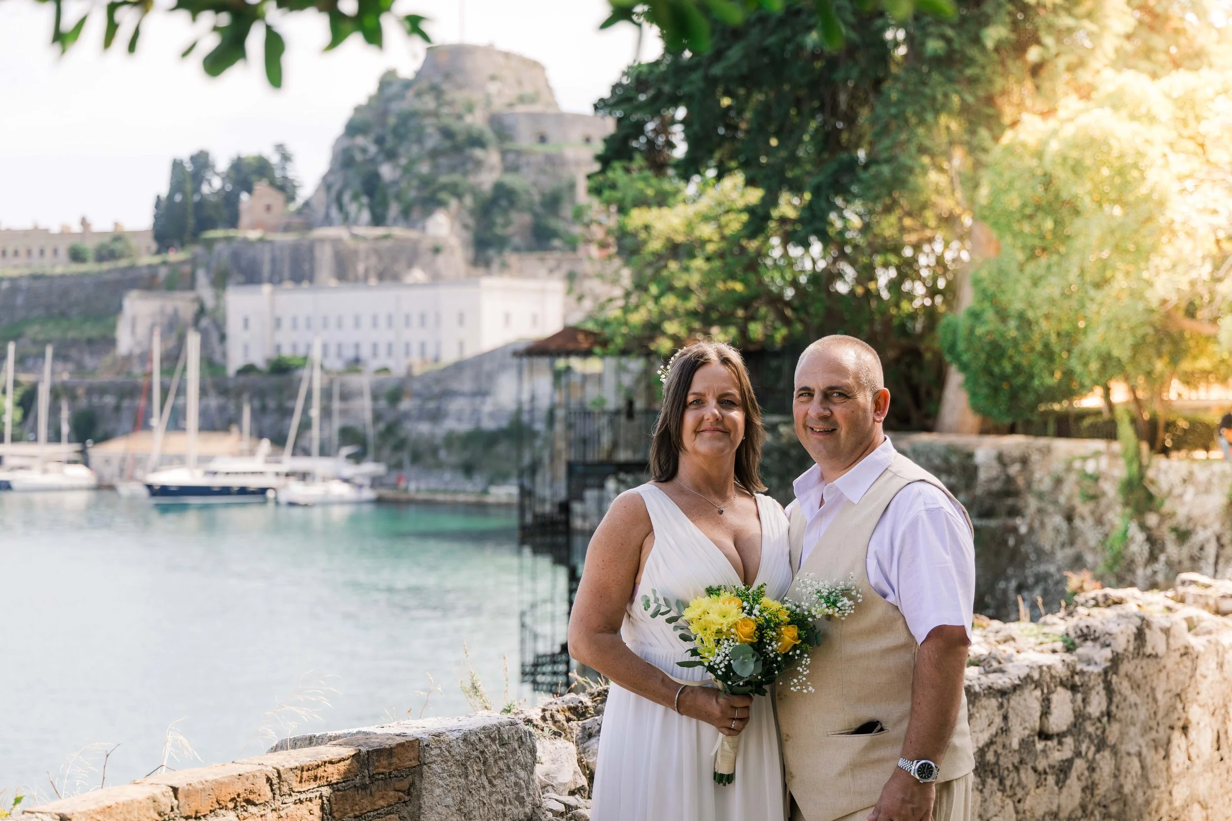 A wedding couple stands outdoors by a stone wall near a lake with sailboats, mountains, and a castle in the background during sunset.