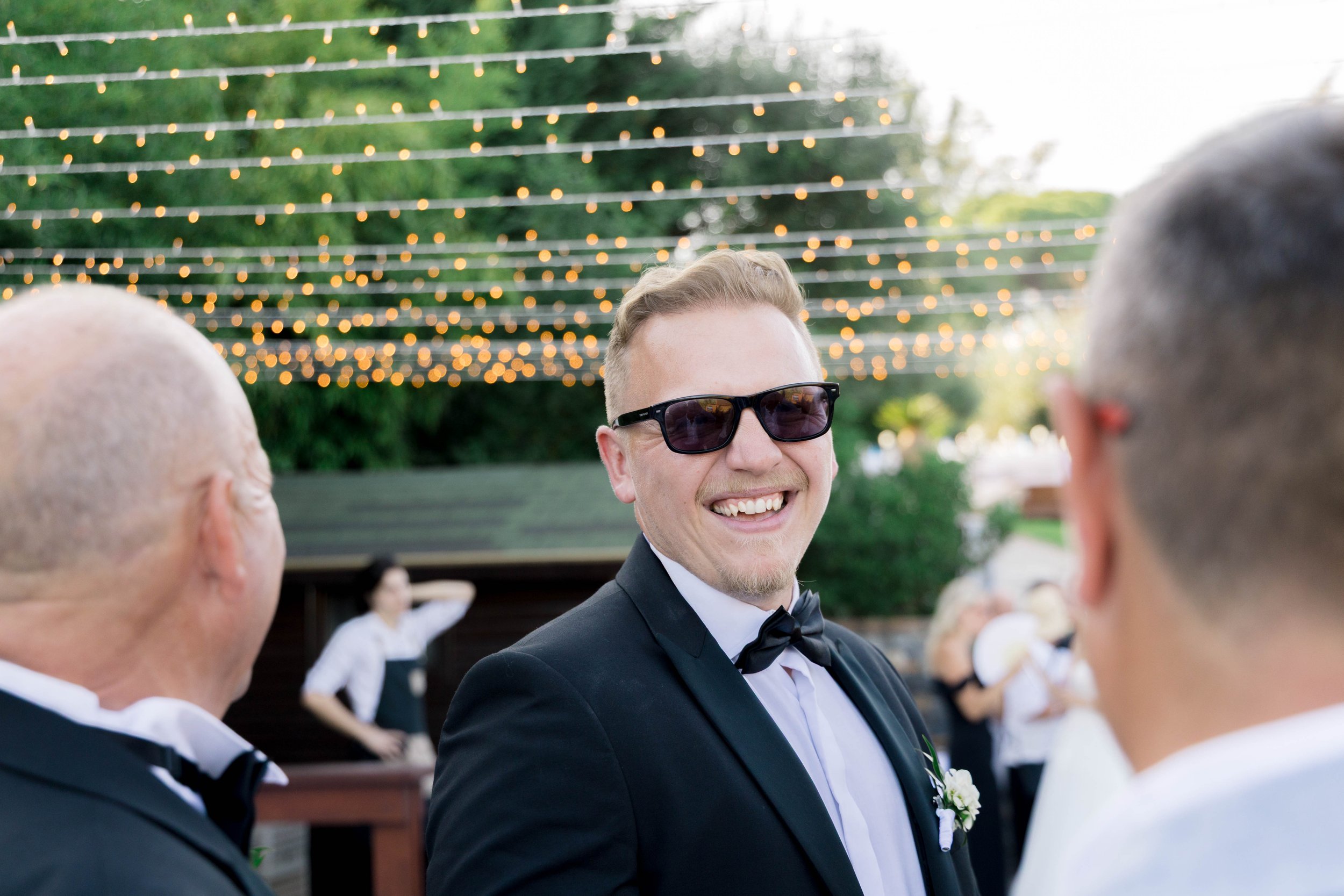 A man in a tuxedo and sunglasses smiling at a wedding reception outdoors with string lights overhead.