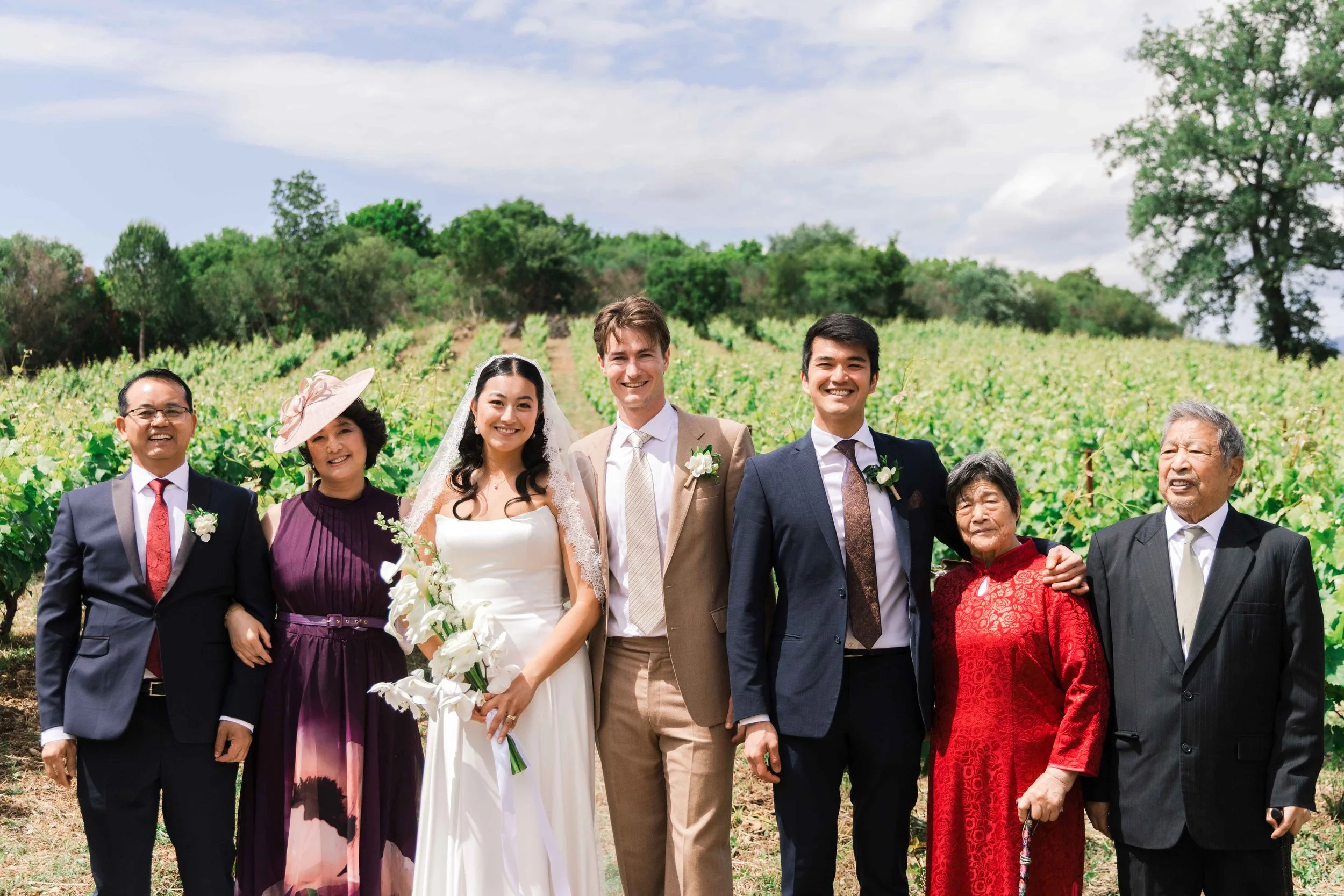 A group of seven people, including a bride and groom, standing together outdoors in a field with green trees and bushes in the background, celebrating a wedding.