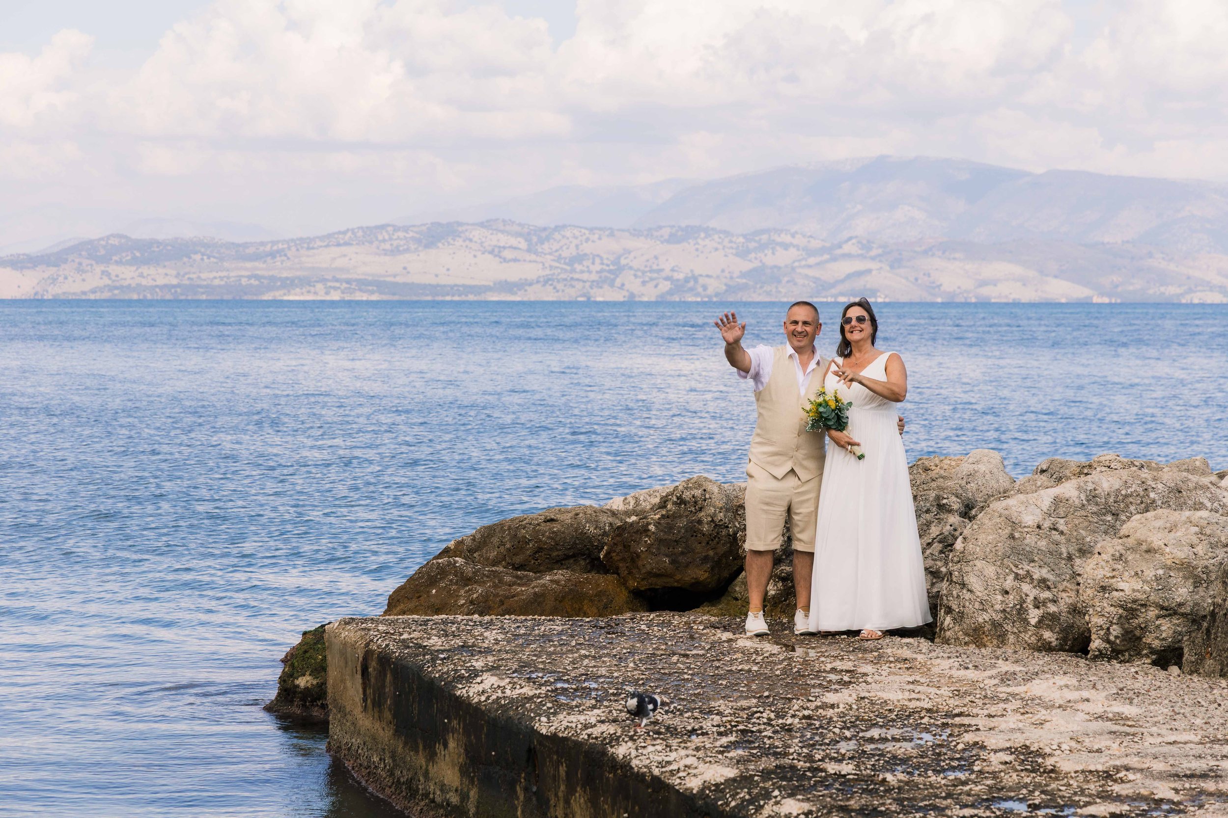 A couple dressed in wedding attire standing on a rocky pier by the water, smiling and waving, with mountains and cloudy sky in the background.