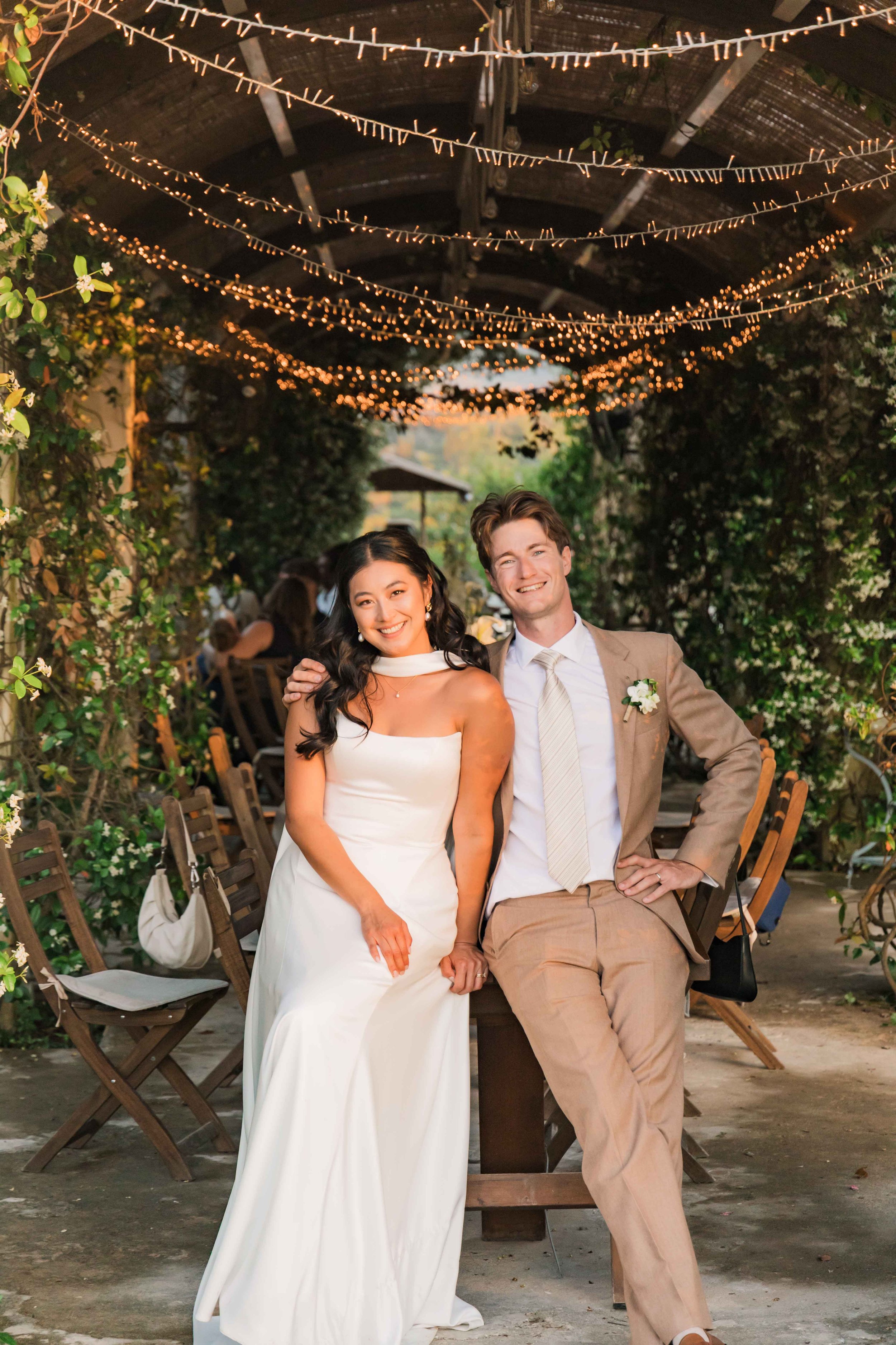 A happy bride and groom sit together during their wedding reception under string lights and greenery, smiling at the camera.