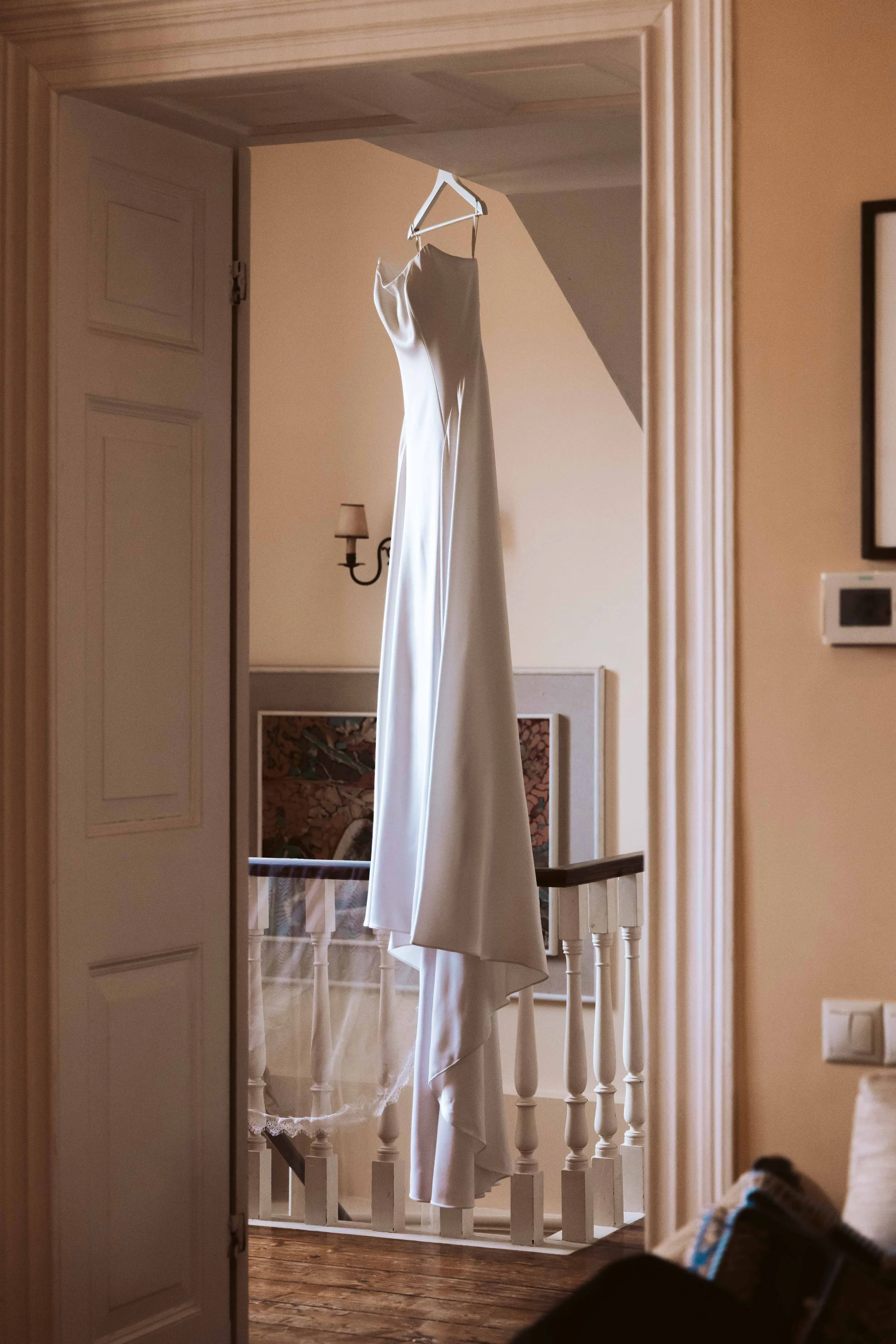 A white wedding dress hangs on a hanger from a doorway, with a staircase and a framed picture in the background.