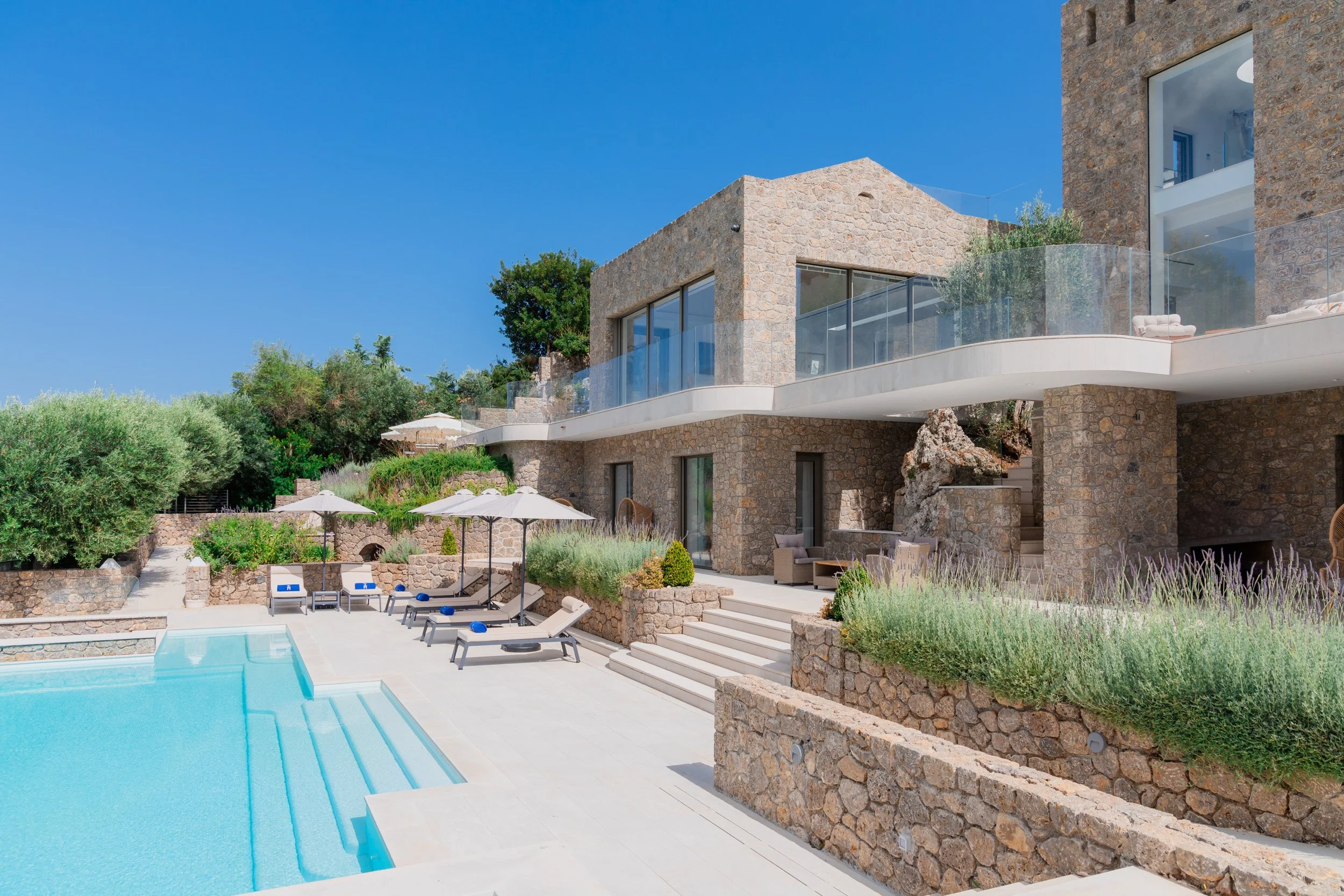 Modern stone house with large glass windows and a balcony overlooking a swimming pool area with lounge chairs and umbrellas, under a clear blue sky.