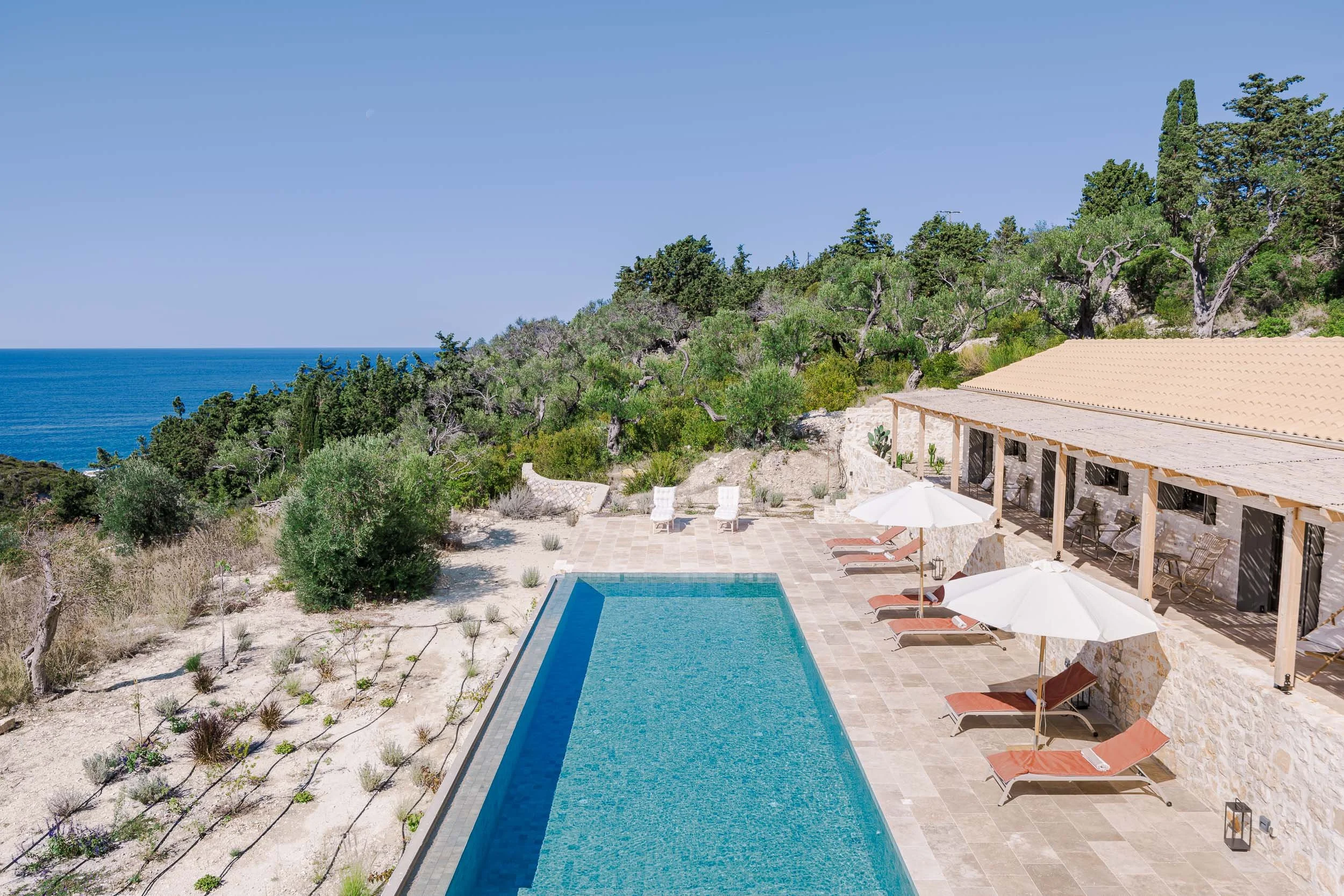A private poolside patio area with lounge chairs, umbrellas, and a view of the ocean and greenery on a sunny day.