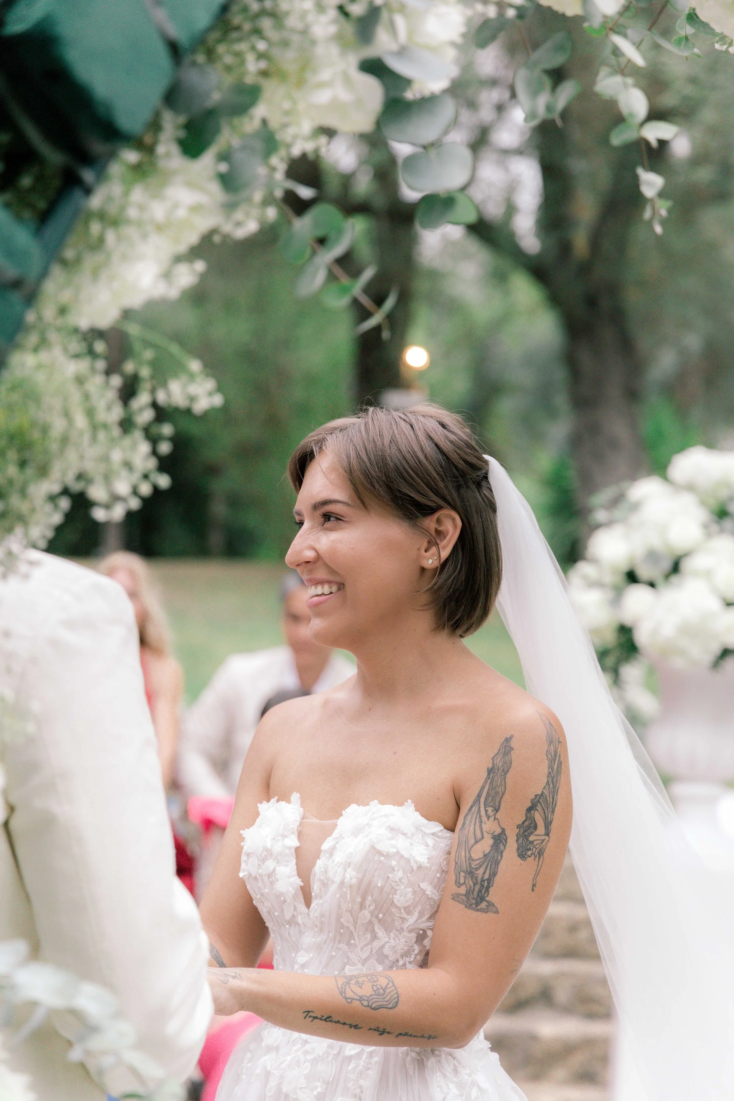 Smiling bride in a strapless white wedding dress with tattoos, participating in an outdoor wedding ceremony with greenery and white floral decorations.