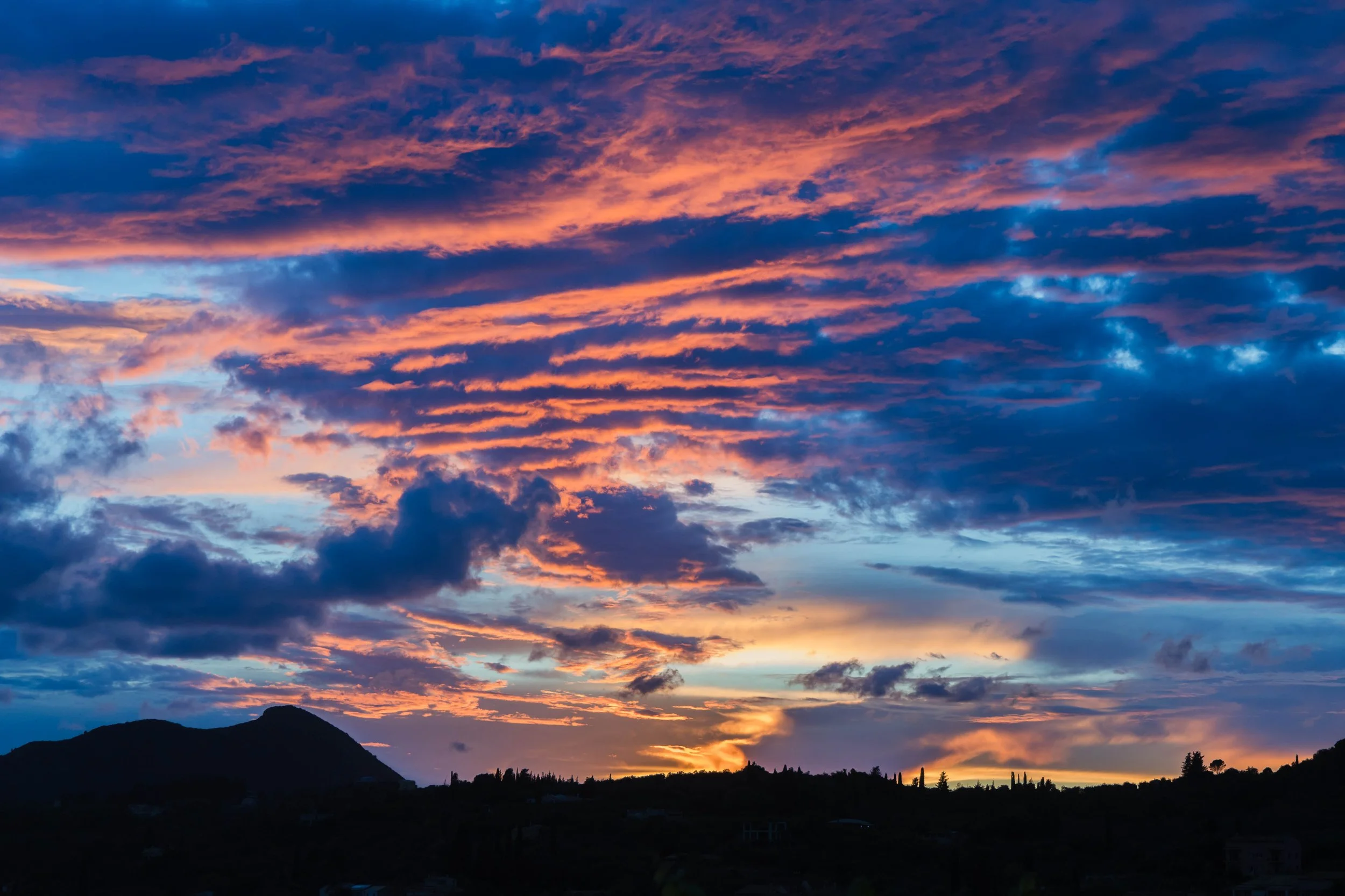 A vibrant sunset over a silhouette of mountains and trees, with colorful clouds in shades of purple, pink, orange, and blue.